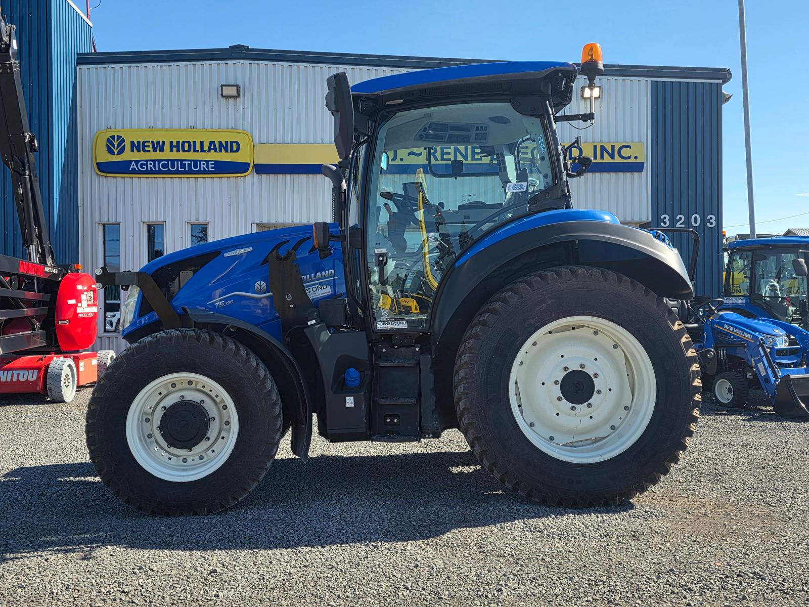 New Holland T5 vu de côté, cabine vitrée, enseigne New Holland Agriculture et bâtiment du concessionnaire en arrière-plan