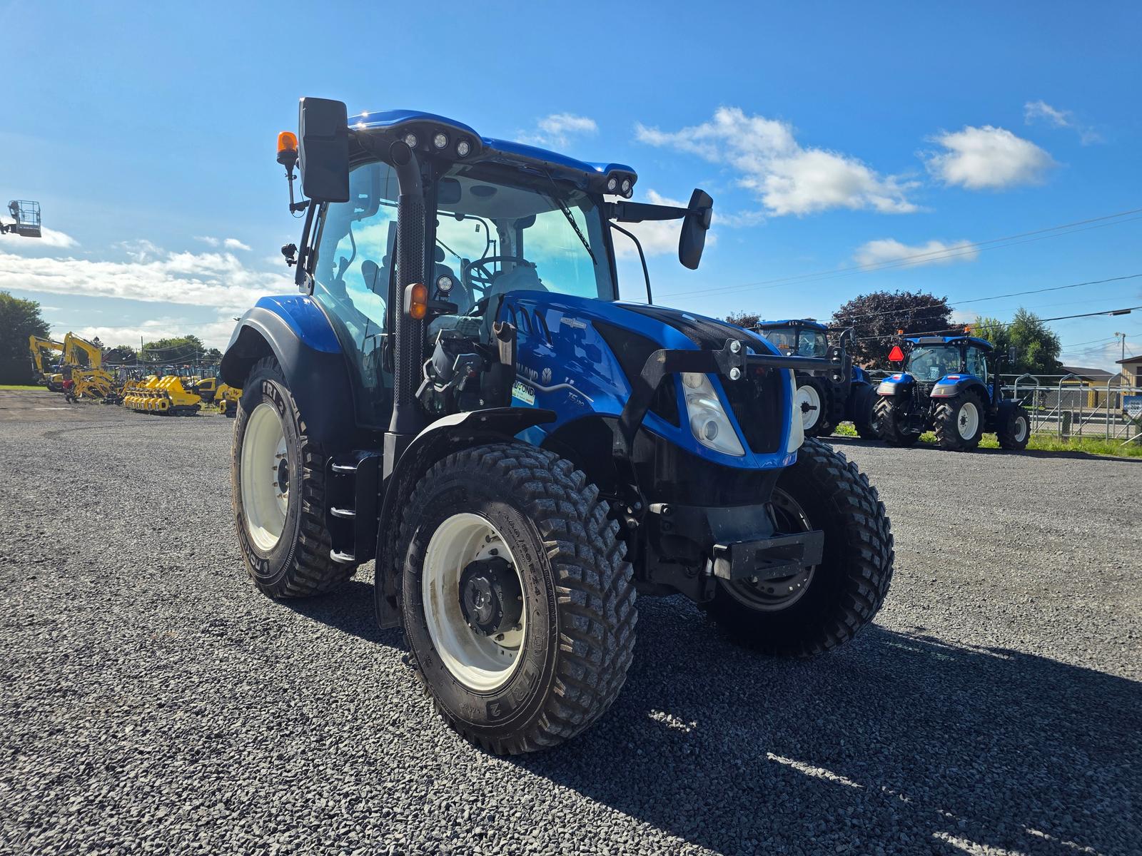 New Holland T5 vu de trois quarts avant, autres tracteurs et machinerie stationnés en arrière-plan