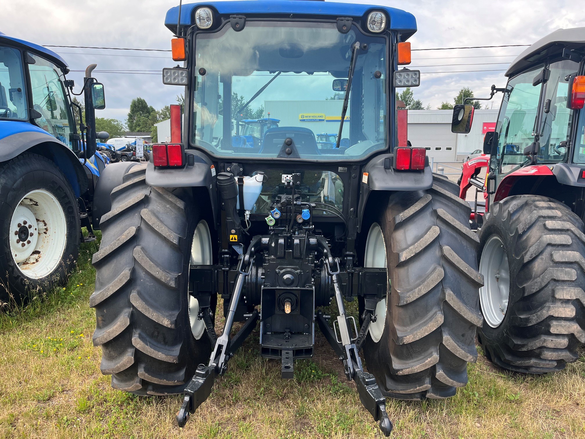 Vue arrière d'un tracteur bleu garé sur l'herbe, flanqué de deux autres tracteurs. L'image montre les gros pneus arrière, les raccords hydrauliques et la cabine du tracteur, avec un ciel nuageux en arrière-plan.
