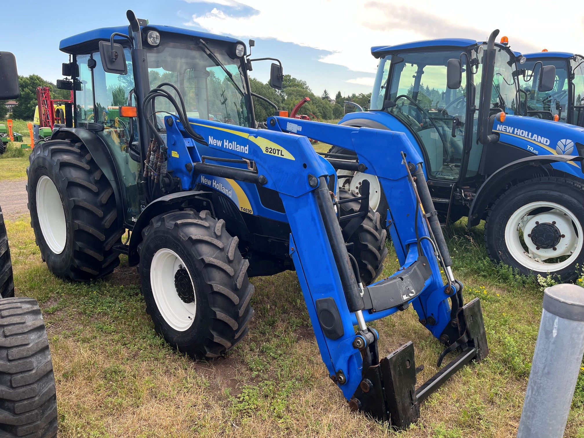 Un tracteur New Holland 220TL bleu équipé d'un chargeur frontal est garé sur l'herbe, à côté d'un autre tracteur similaire, sous un ciel partiellement nuageux.