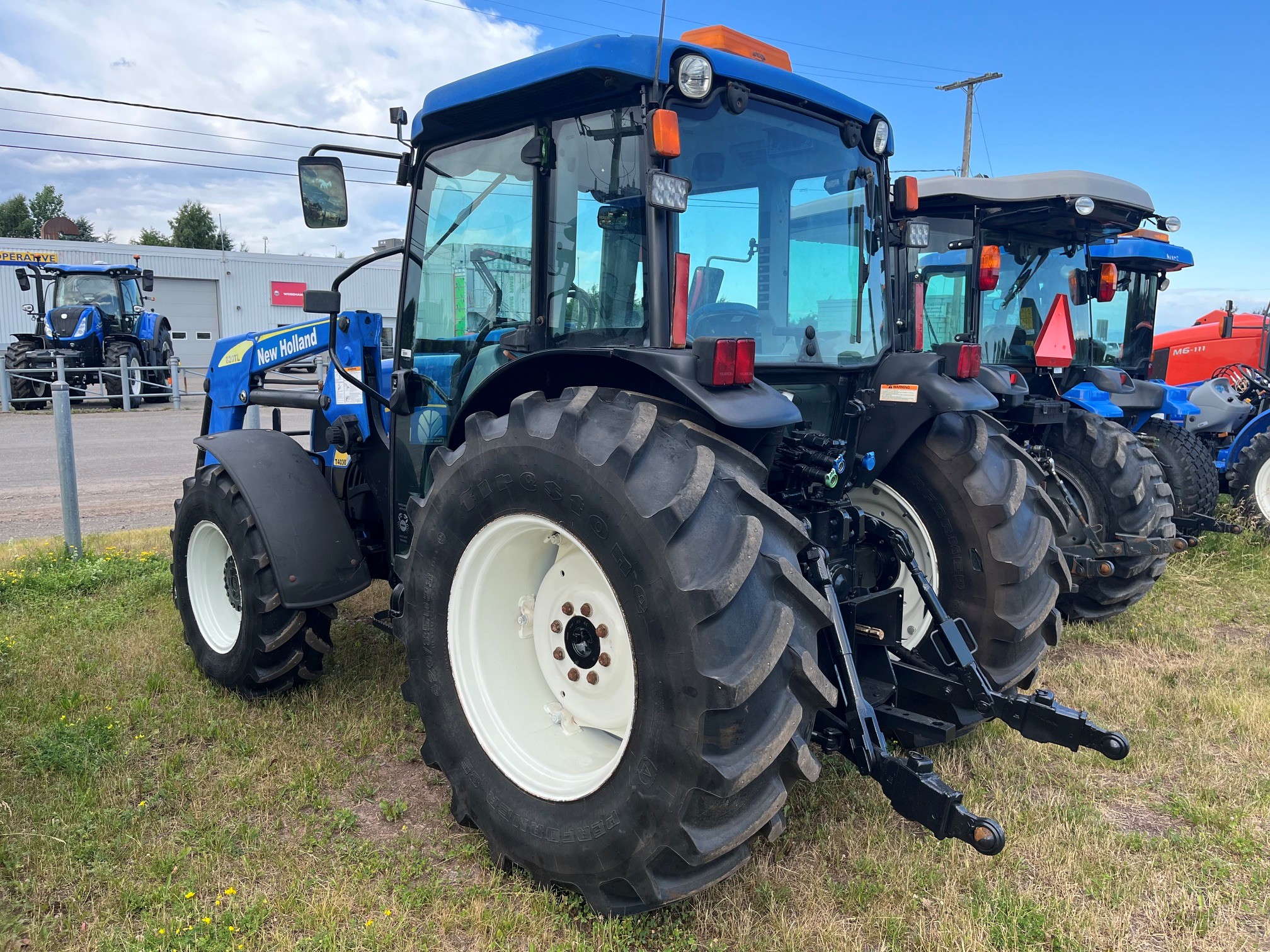 Un tracteur New Holland bleu est garé sur l'herbe, près d'autres tracteurs ; il est équipé de gros pneus arrière et d'une cabine, sous un ciel partiellement nuageux.