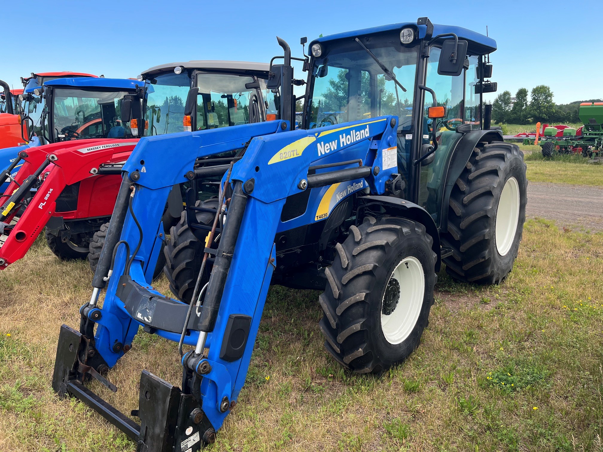 Un tracteur New Holland bleu équipé d'un chargeur frontal est garé sur l'herbe, à côté d'autres tracteurs rouges et verts, sous un ciel bleu limpide.