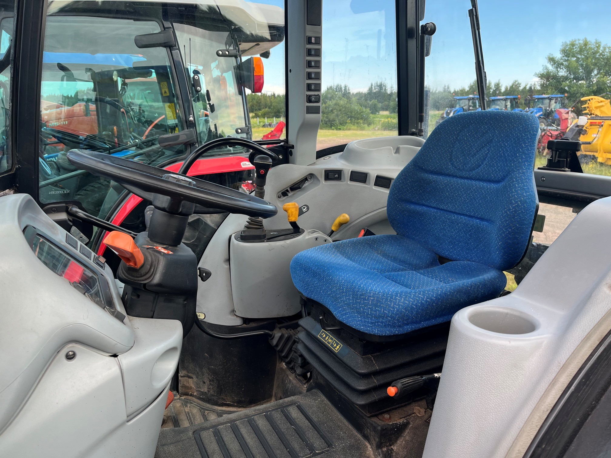 Intérieur d'une cabine de tracteur montrant un siège conducteur bleu, un volant, des tableaux de bord et des leviers de vitesses. Les vitres de la cabine offrent une vue sur des machines à l'extérieur et de la verdure en arrière-plan.