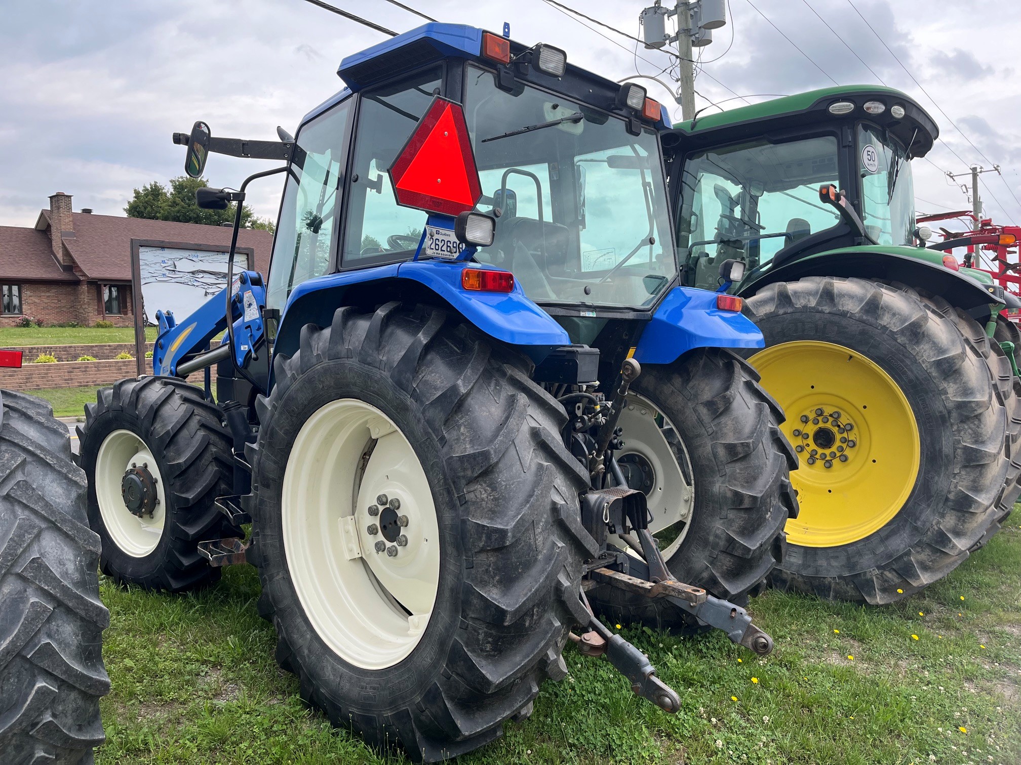 Un tracteur bleu équipé de gros pneus arrière est garé sur l'herbe, à côté d'un tracteur vert. Un triangle réfléchissant rouge se trouve à l'arrière, et un bâtiment au toit marron est visible à l'arrière-plan.