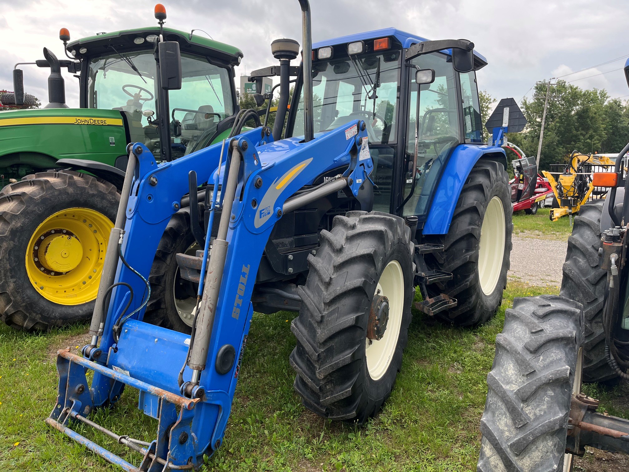 Un tracteur bleu équipé d'un chargeur frontal est garé sur l'herbe, à côté d'un tracteur John Deere vert et d'un autre véhicule. On aperçoit des arbres et un ciel nuageux en arrière-plan.