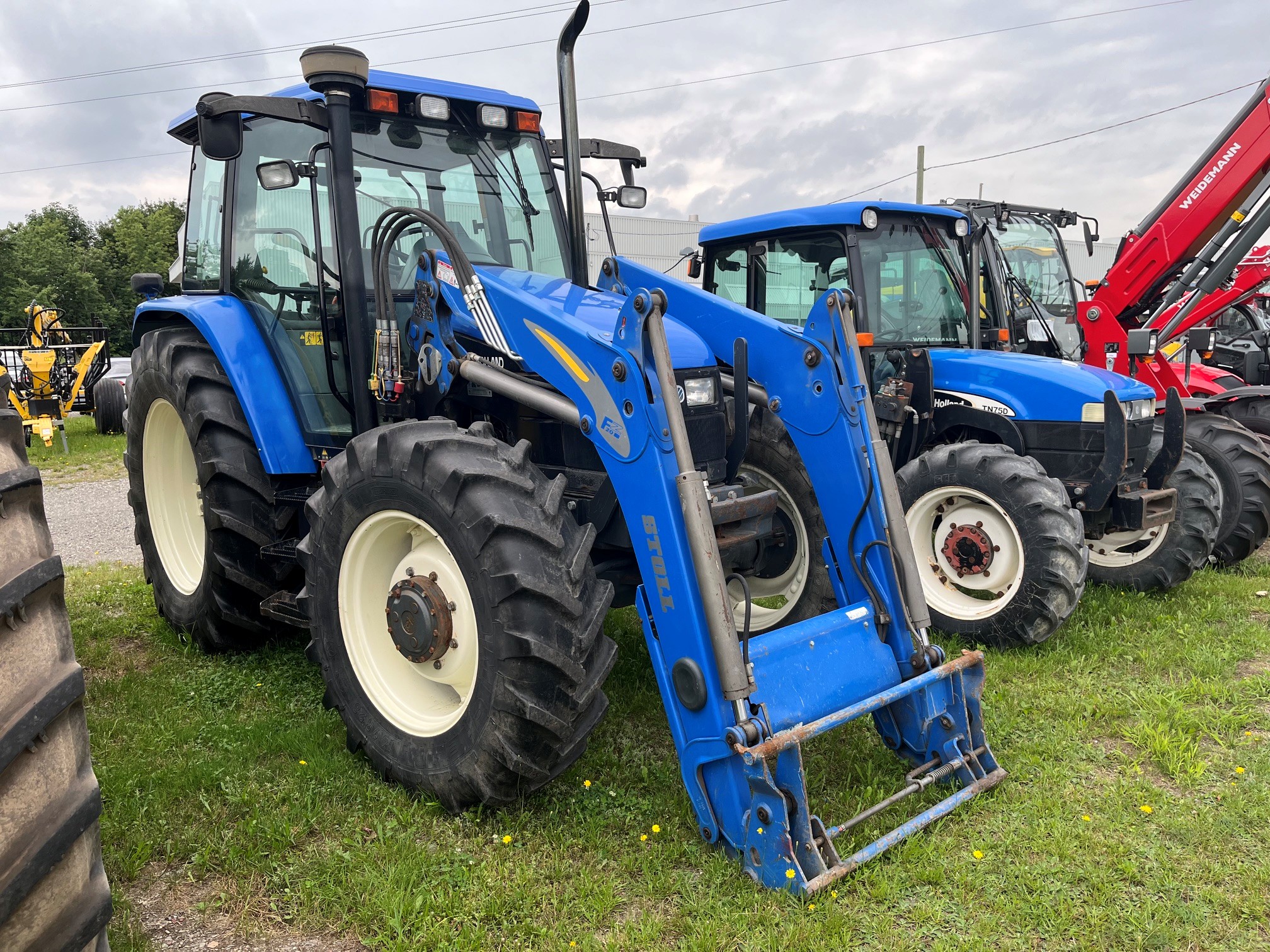 Deux tracteurs bleus équipés de gros pneus sont garés sur l'herbe. Le tracteur du premier plan est équipé d'un chargeur frontal. Un autre tracteur et d'autres engins sont visibles à l'arrière-plan, sous un ciel nuageux.