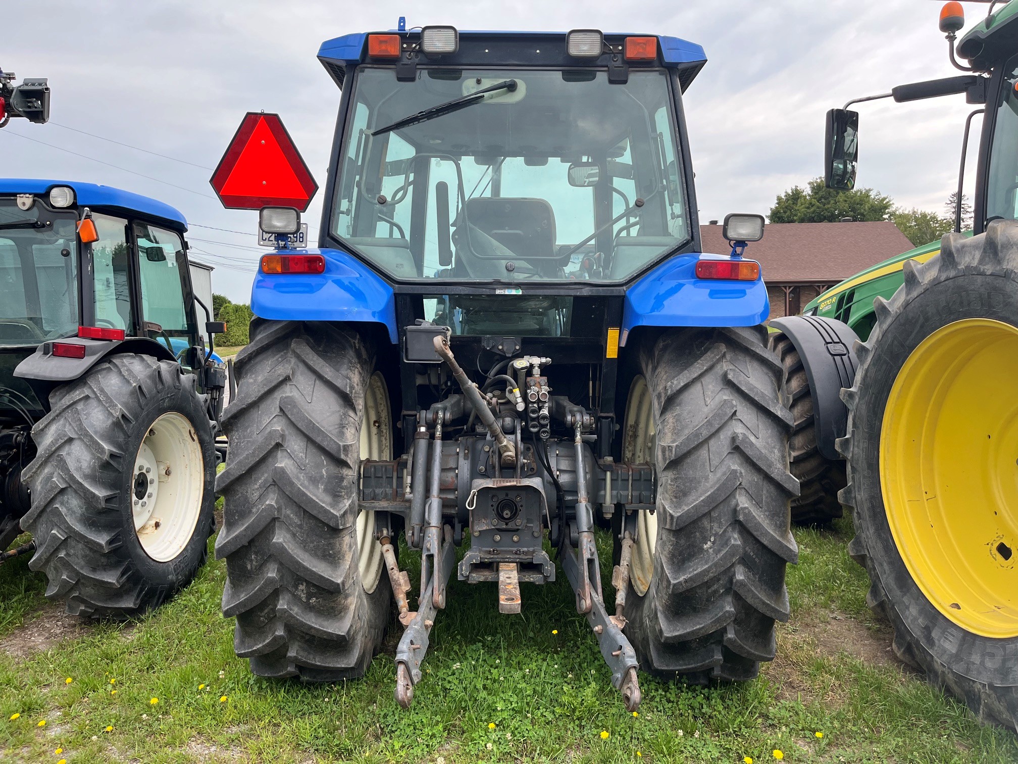 Un tracteur bleu vu de l'arrière, sur lequel on distingue de gros pneus, un triangle réfléchissant et des dispositifs d'attelage. D'autres tracteurs sont visibles de chaque côté, garés sur l'herbe sous un ciel nuageux.