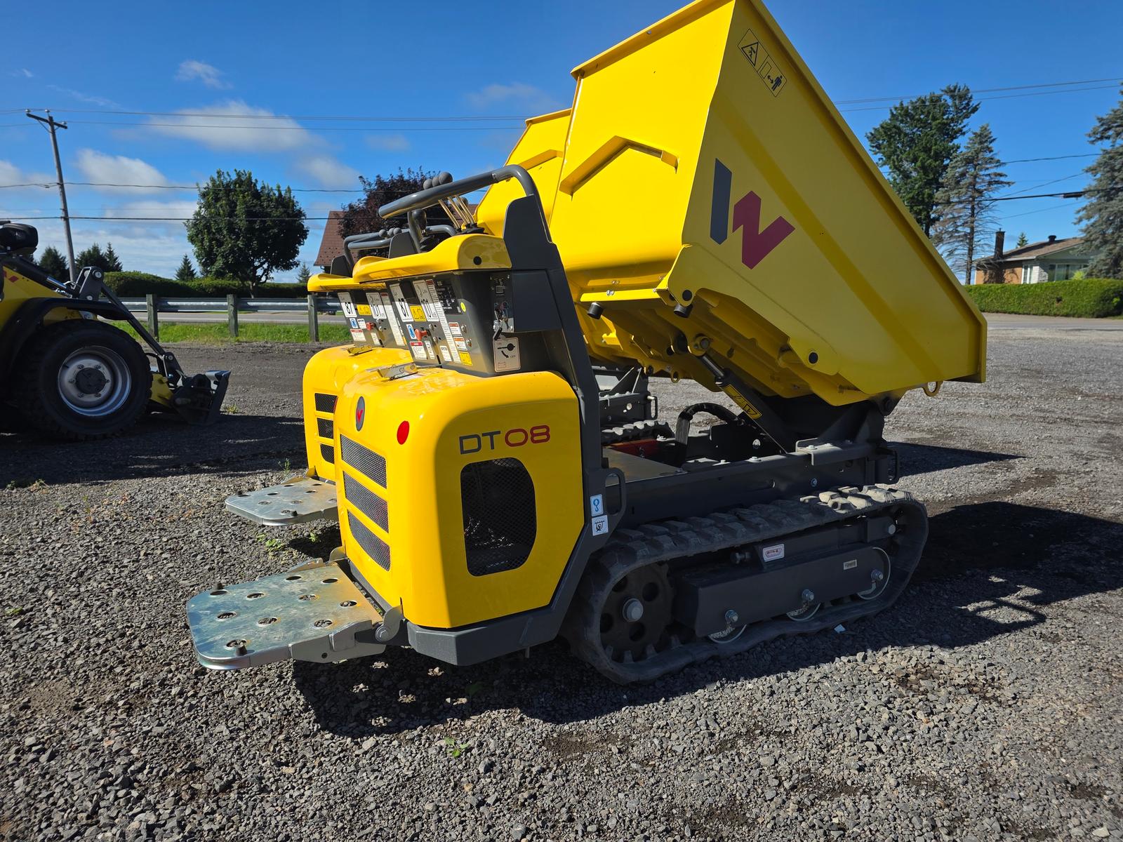Wacker Neuson DT08 vu de côté, benne relevée à l'avant, plaques d'accès au sol abaissées, bâtiment du concessionnaire en arrière-plan