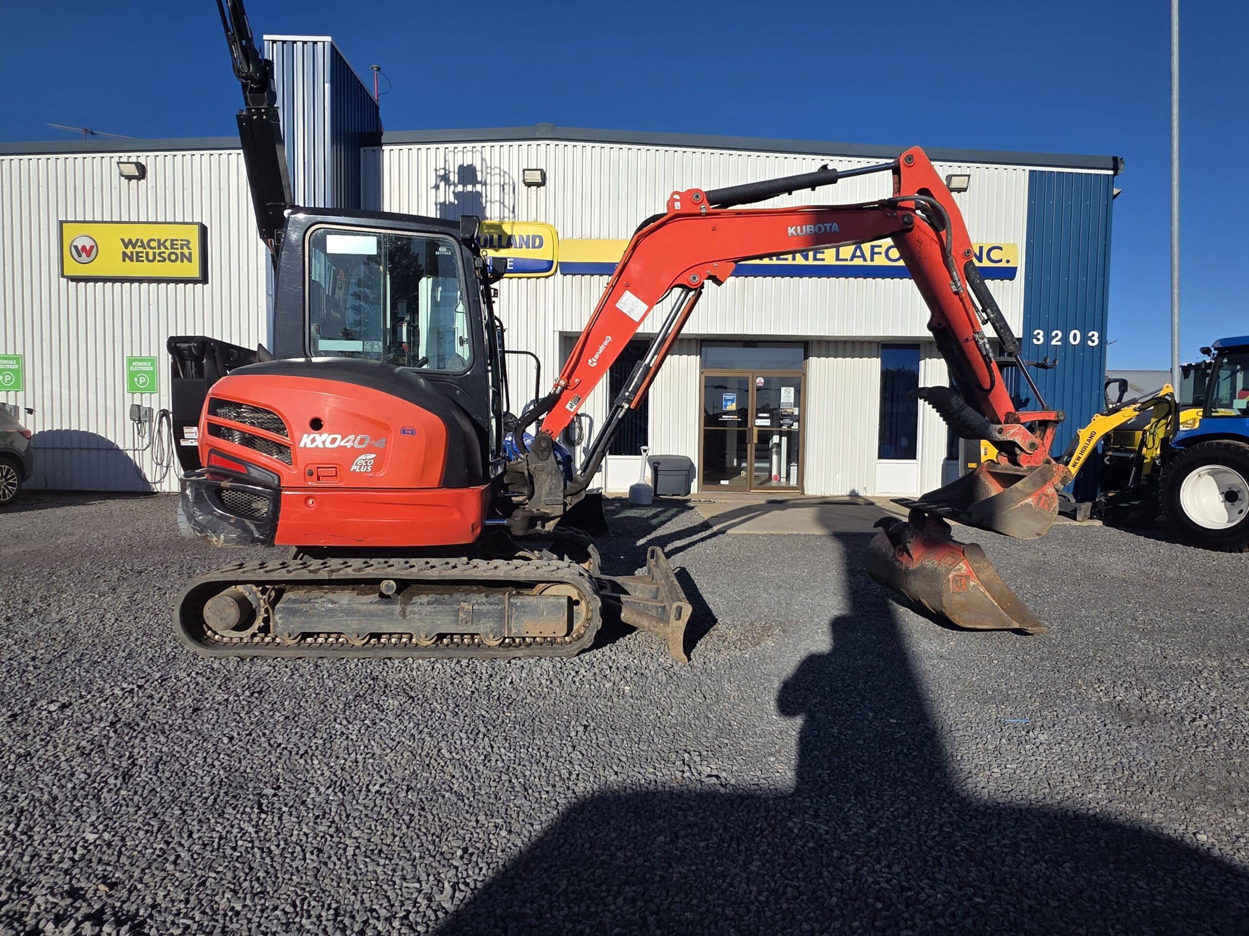 Une petite pelleteuse Kubota KX040-4 rouge garée sur un terrain en gravier devant un bâtiment commercial.