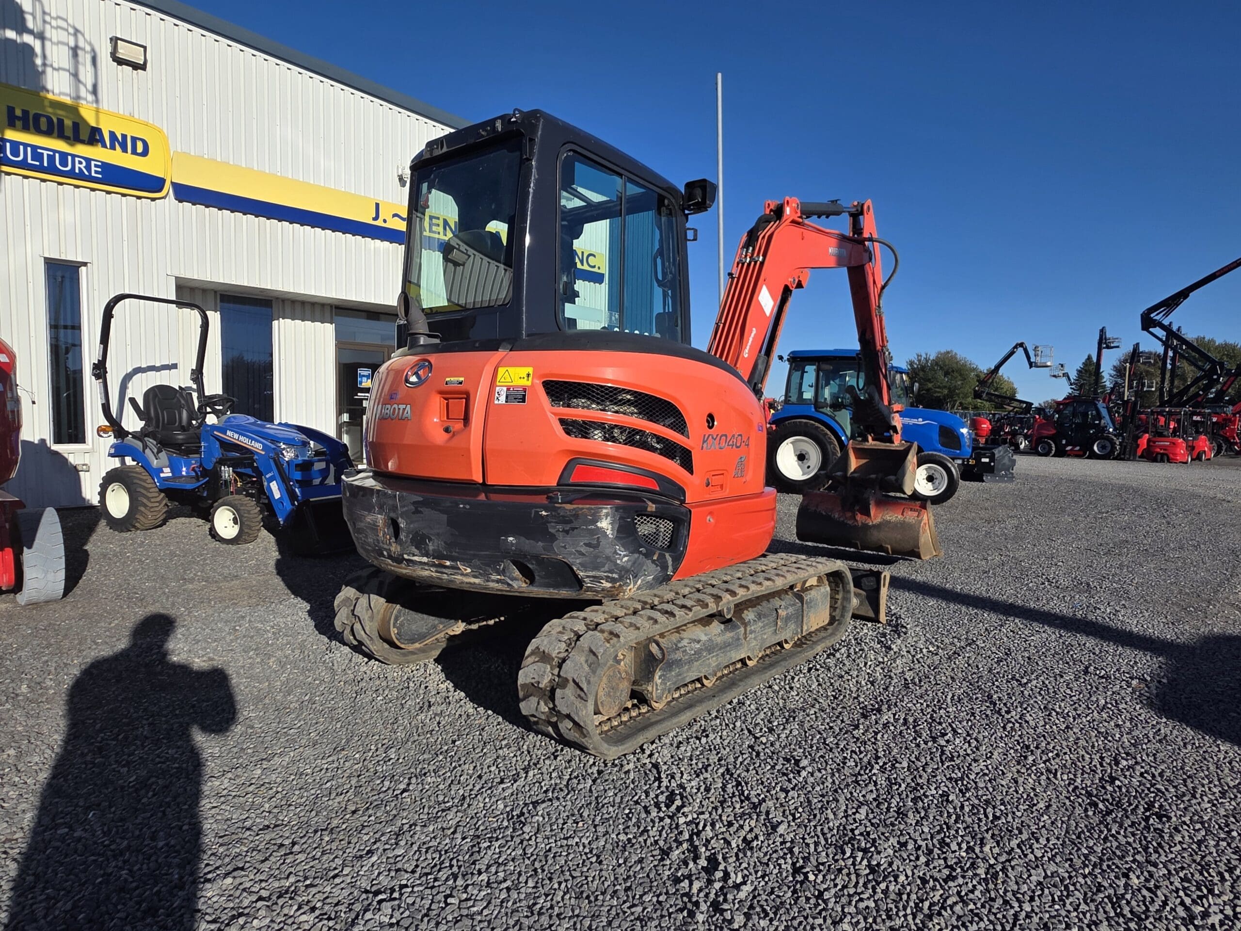 Une mini-pelle Kubota de couleur orange garée sur un terrain en gravier devant un bâtiment, avec d'autres machines agricoles visibles à l'arrière-plan.