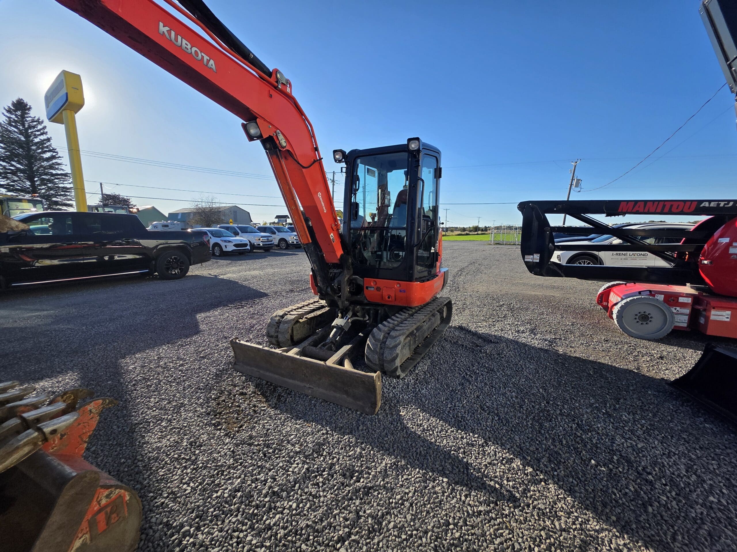 Une mini-pelle Kubota rouge était garée sur un terrain en gravier, à côté d'un pick-up noir et d'une nacelle Manitou.