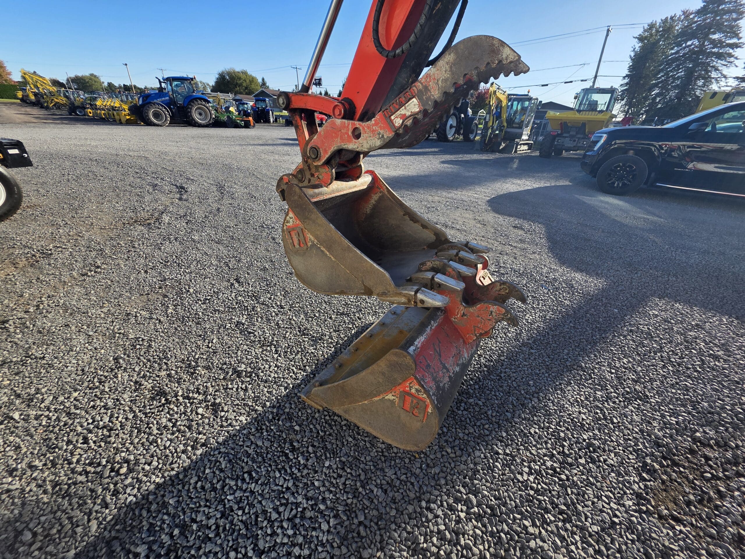Gros plan sur le godet rouge d'une pelleteuse hydraulique suspendu au-dessus d'un terrain en gravier où sont garés divers tracteurs et engins de chantier.