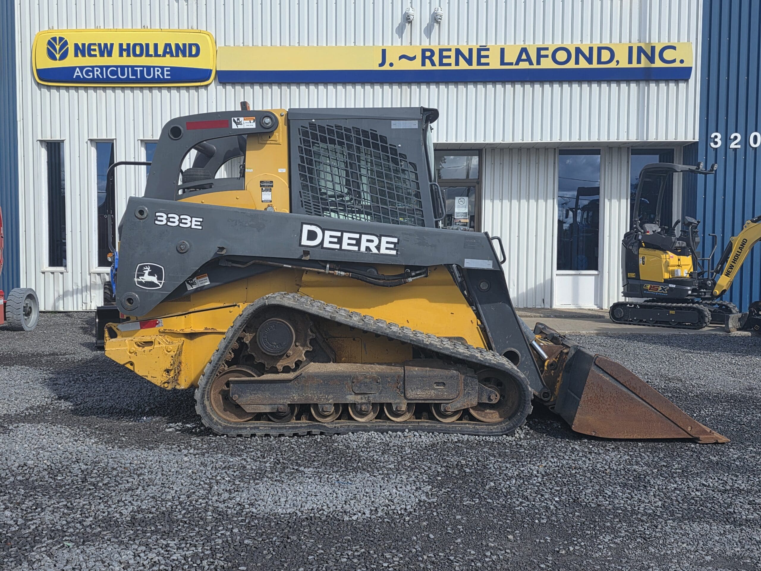 Une chargeuse compacte sur chenilles Yellow Deere 333E garée devant une concession New Holland.