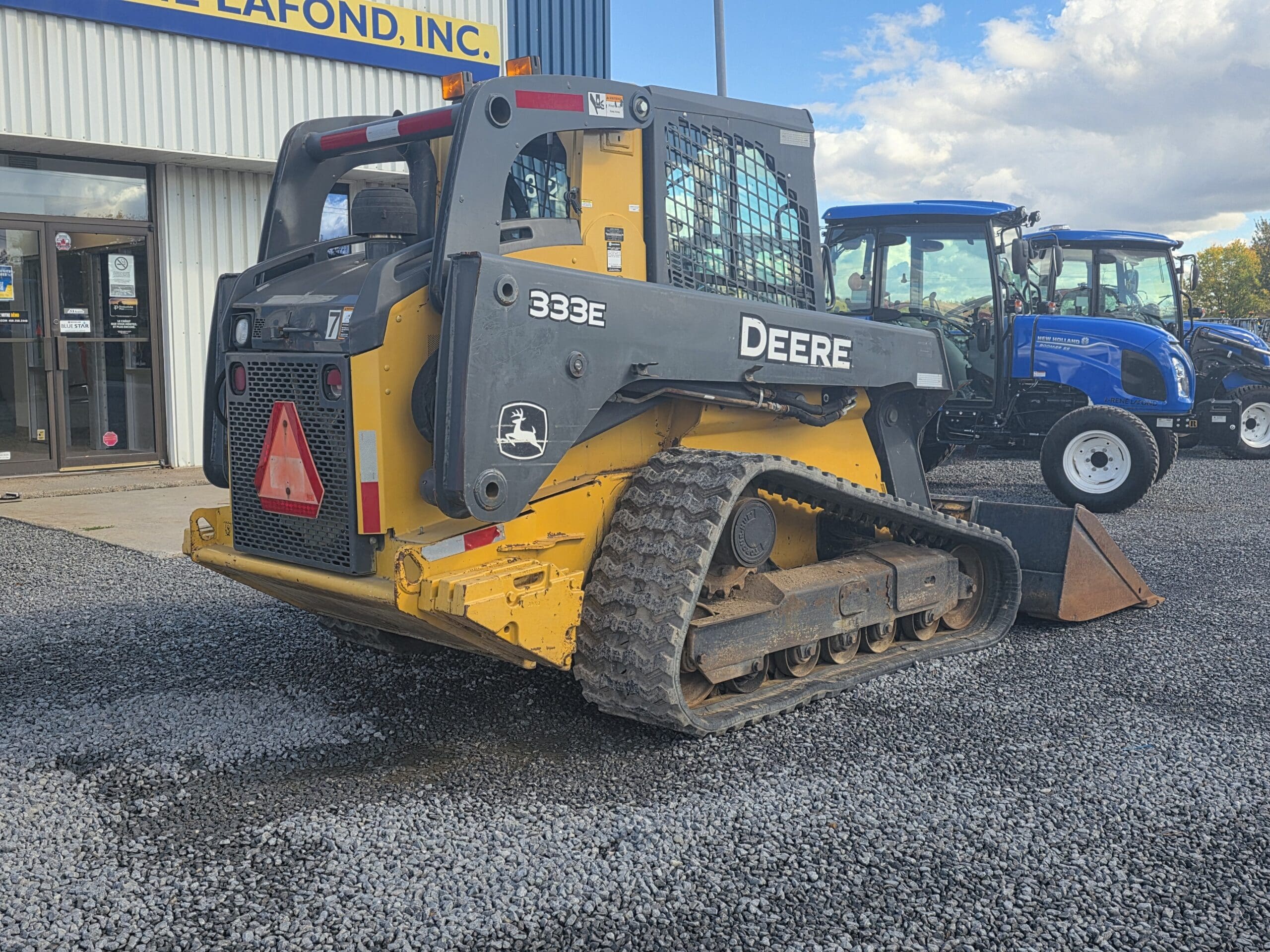 Une chargeuse compacte sur chenilles John Deere 333E jaune, garée sur un terrain en gravier à côté d'un tracteur bleu.
