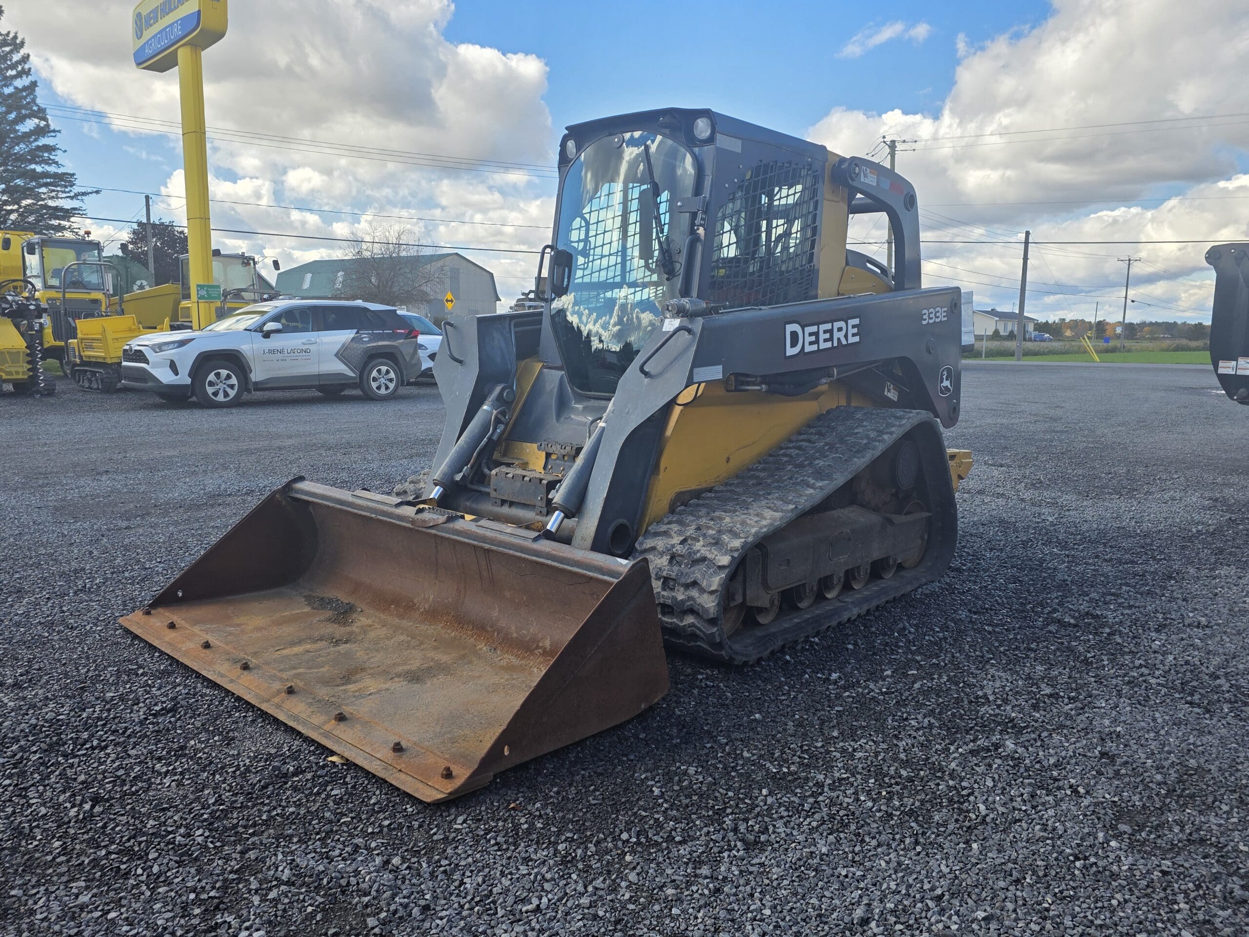 Chargeuse compacte sur chenilles John Deere 333E équipée d'un godet métallique, stationnée sur un terrain en gravier.