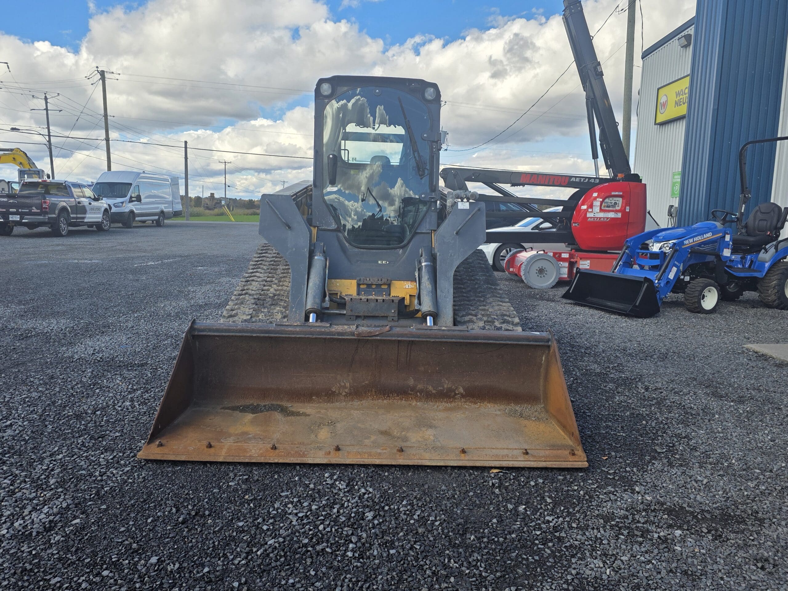 Une chargeuse compacte à chenilles noire garée sur un terrain en gravier, avec un tracteur compact New Holland bleu à sa droite.
