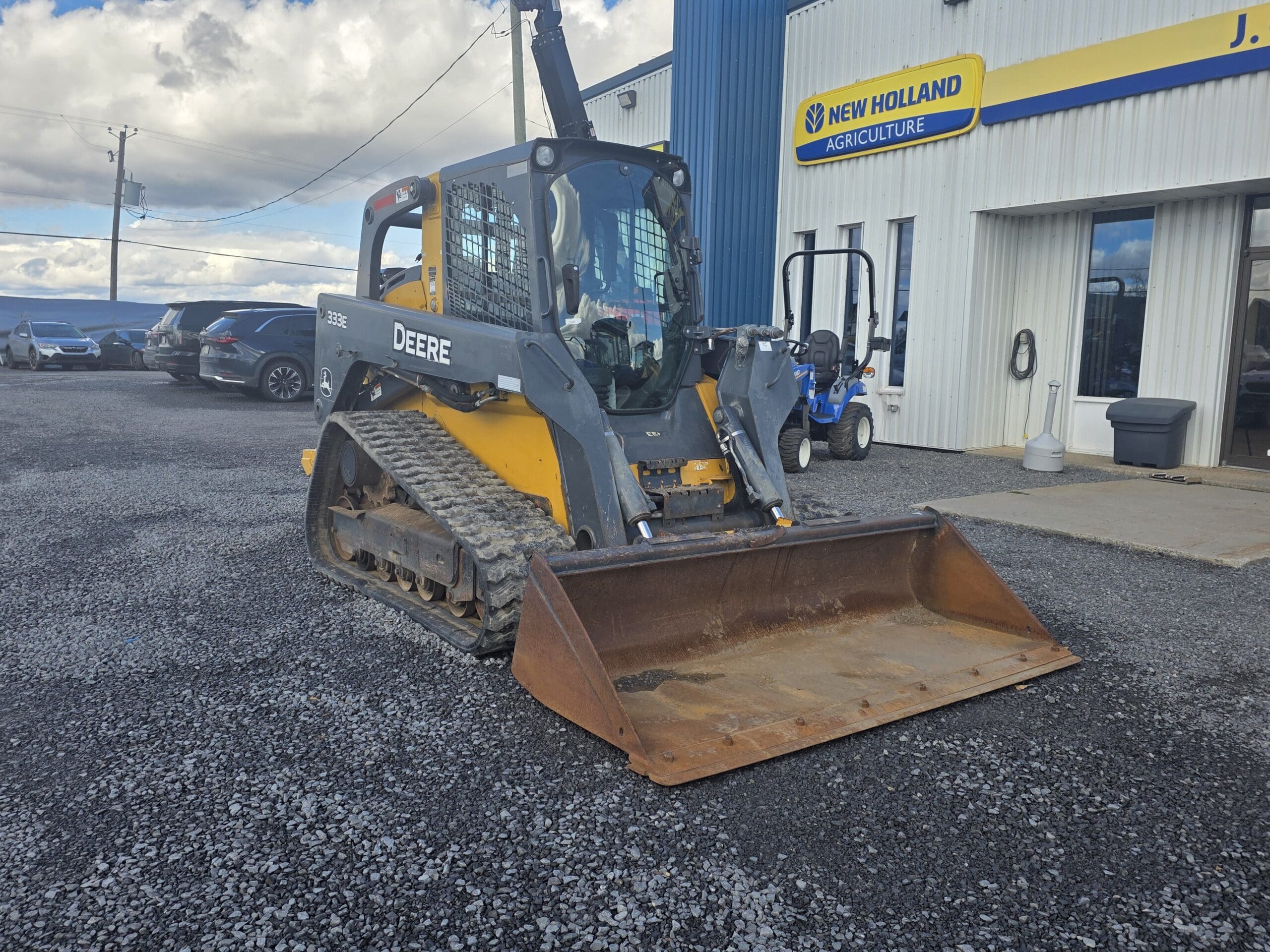 Une chargeuse compacte sur chenilles John Deere 333E, de couleur jaune et grise, équipée d'un godet, garée devant une concession New Holland.