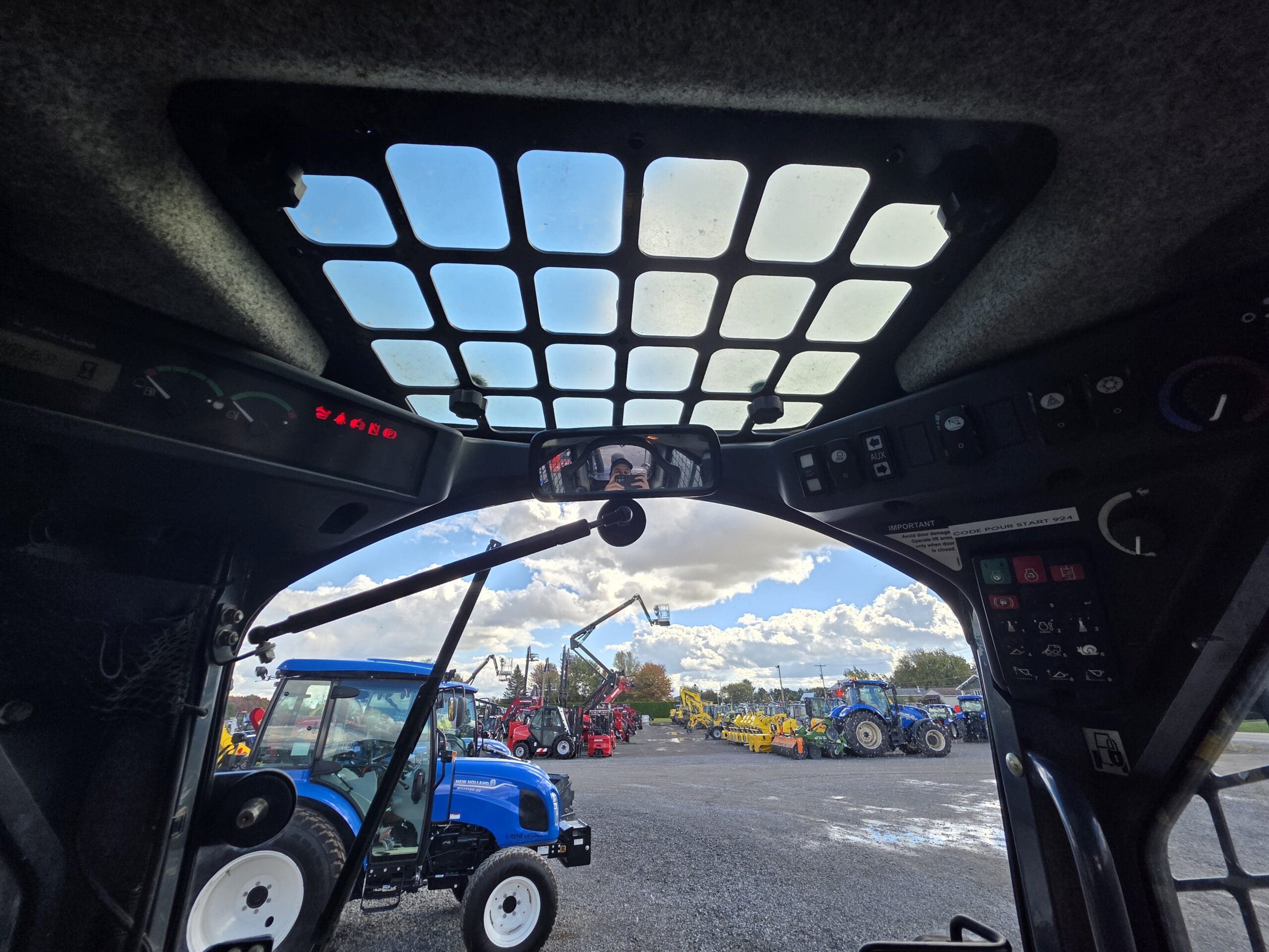 Vue depuis l'intérieur d'une chargeuse compacte donnant sur un terrain où sont entreposés divers tracteurs et matériels agricoles.