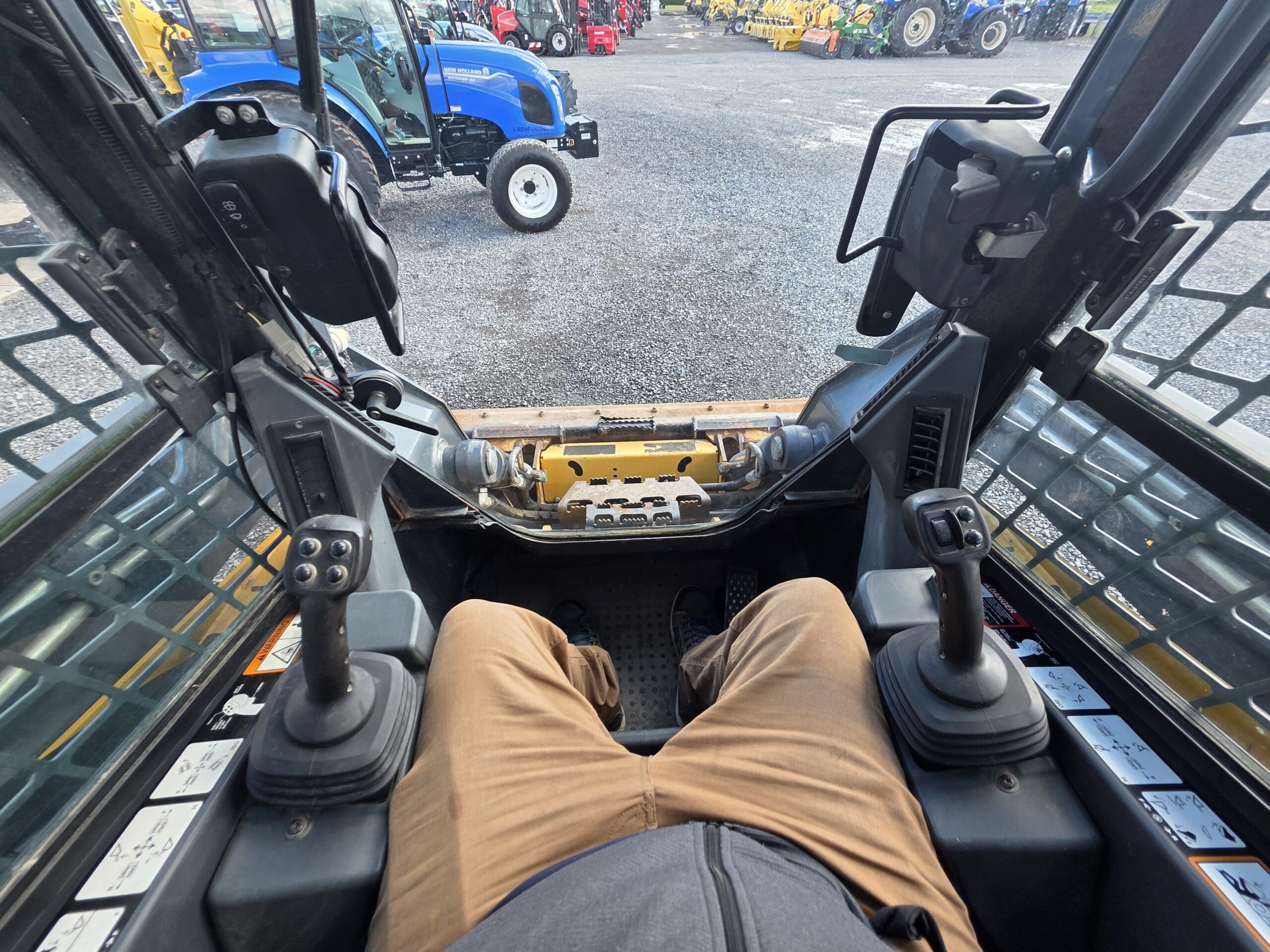 Vue depuis l'intérieur de la cabine d'une chargeuse compacte donnant sur un terrain en gravier où sont garés des tracteurs.