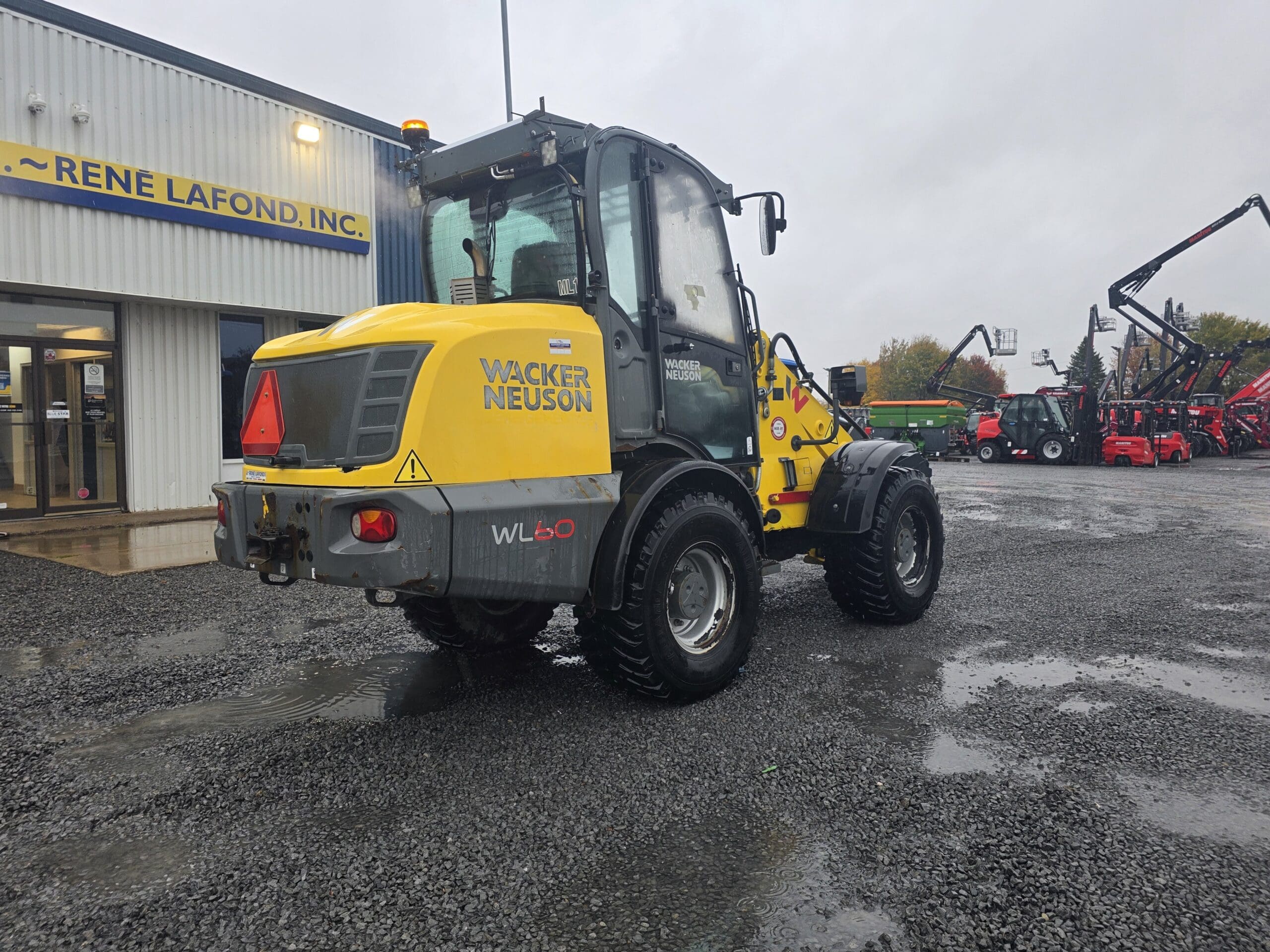 Une chargeuse sur pneus Wacker Neuson WL60 de couleur jaune, garée sur un parking en gravier devant un bâtiment de la société René Lafond, Inc.