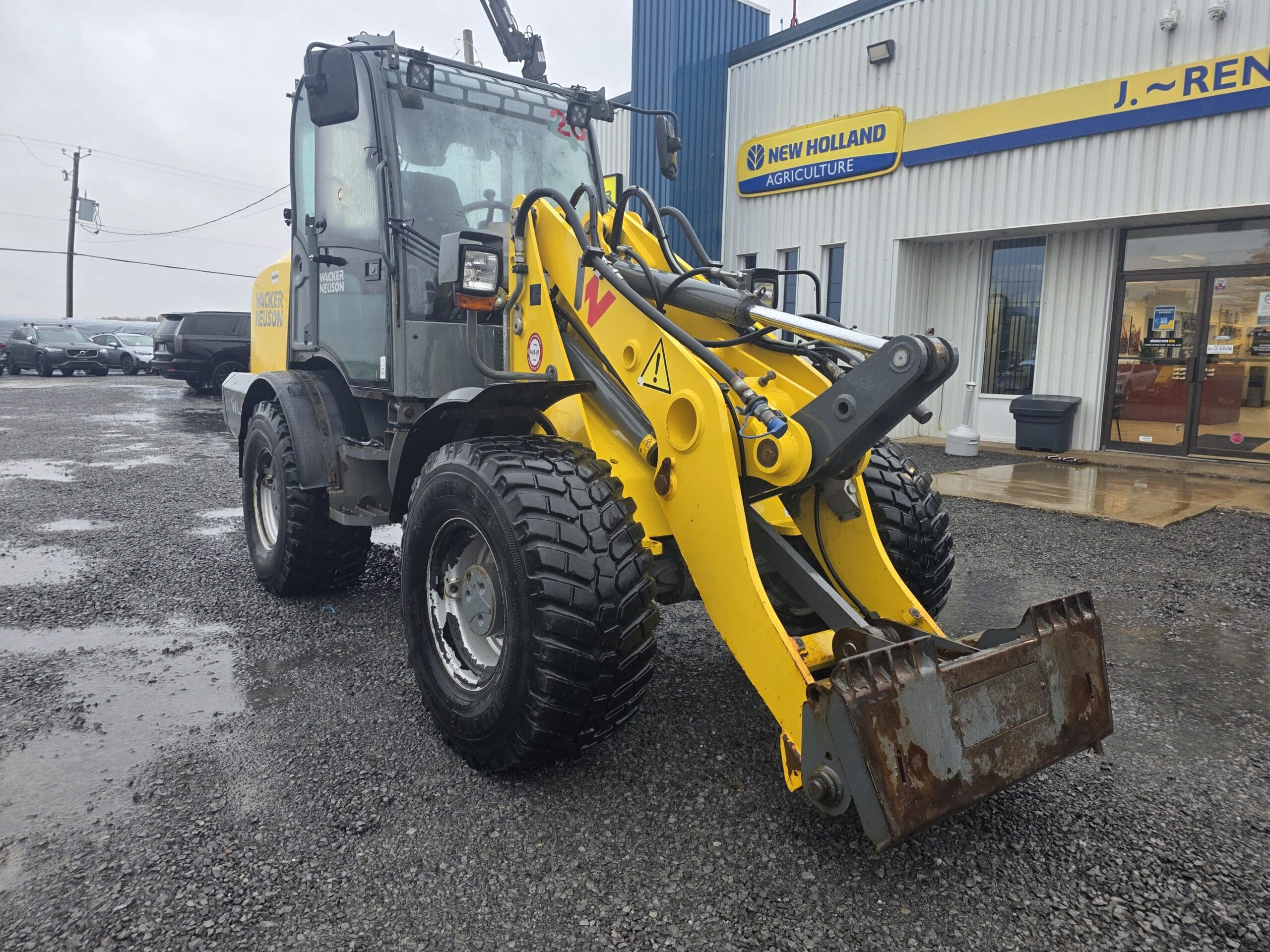 Une chargeuse sur pneus jaune de marque Wacker Neuson, équipée d'un godet avant, est garée sur un parking en gravier devant un concessionnaire New Holland Agriculture.