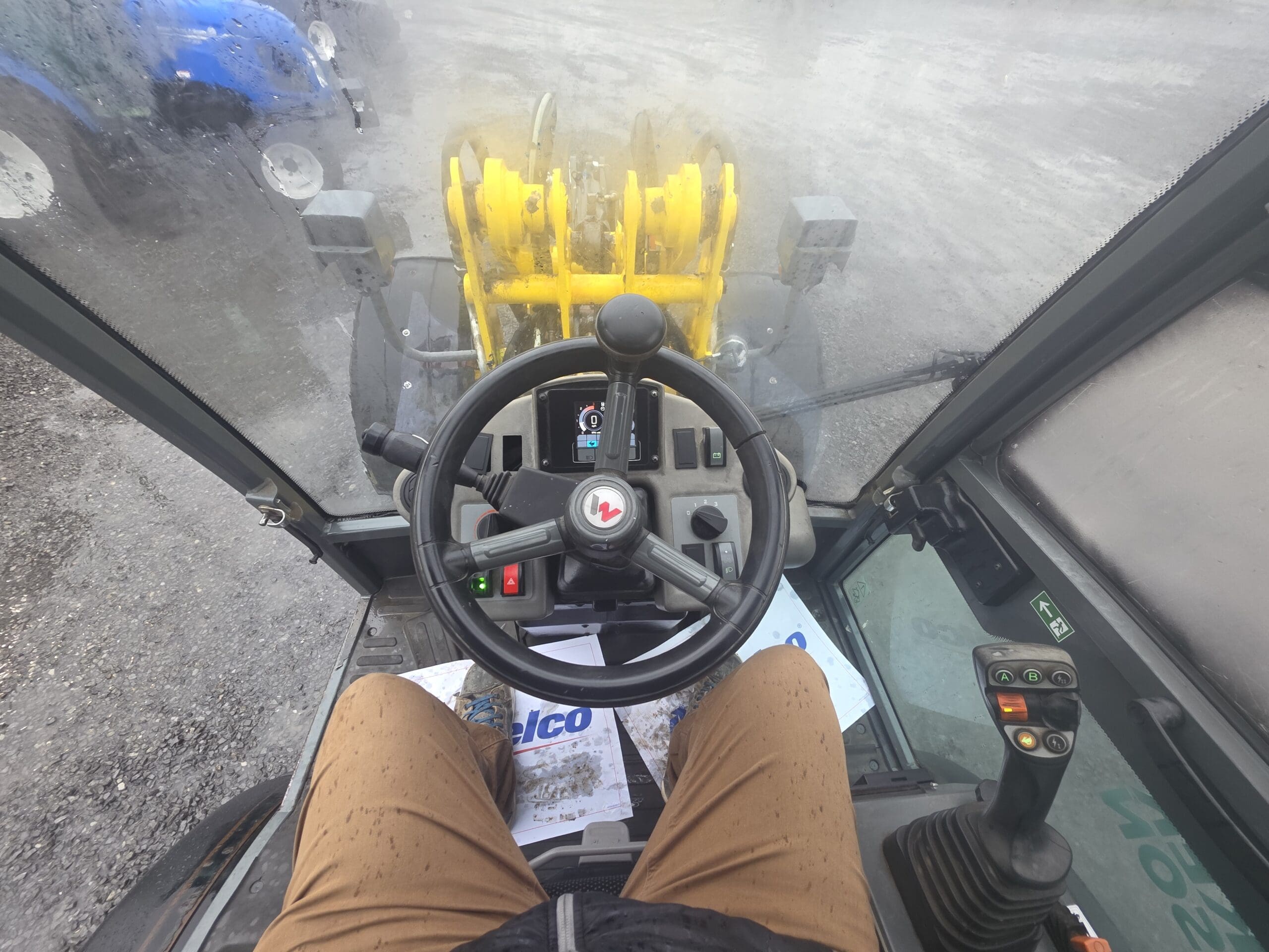 Vue depuis la cabine d'un tracteur montrant le volant et les commandes, avec un équipement jaune visible à travers le pare-brise avant.