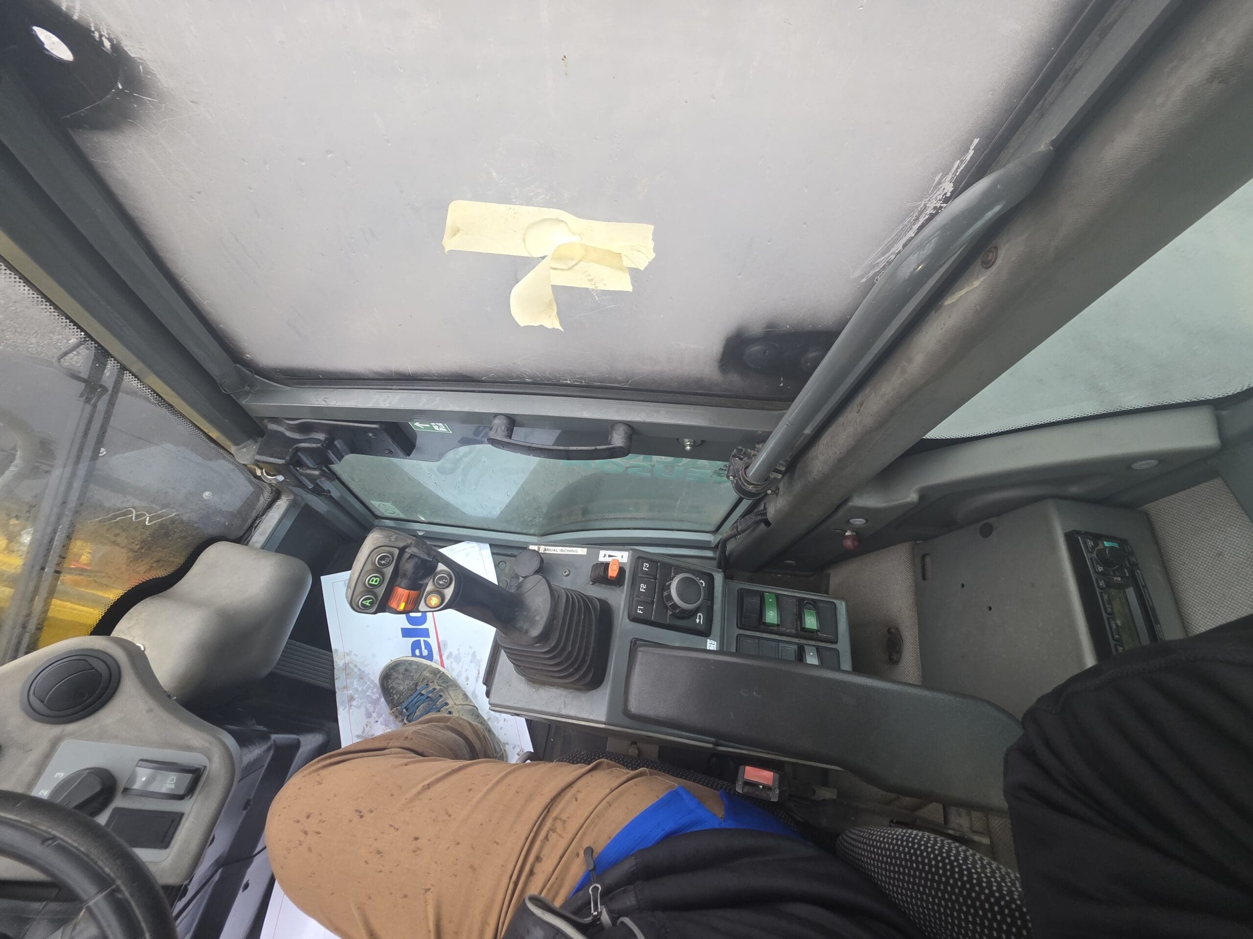 Vue intérieure depuis le siège du conducteur d'un engin de chantier, montrant un joystick de commande, des boutons et les jambes de l'opérateur.
