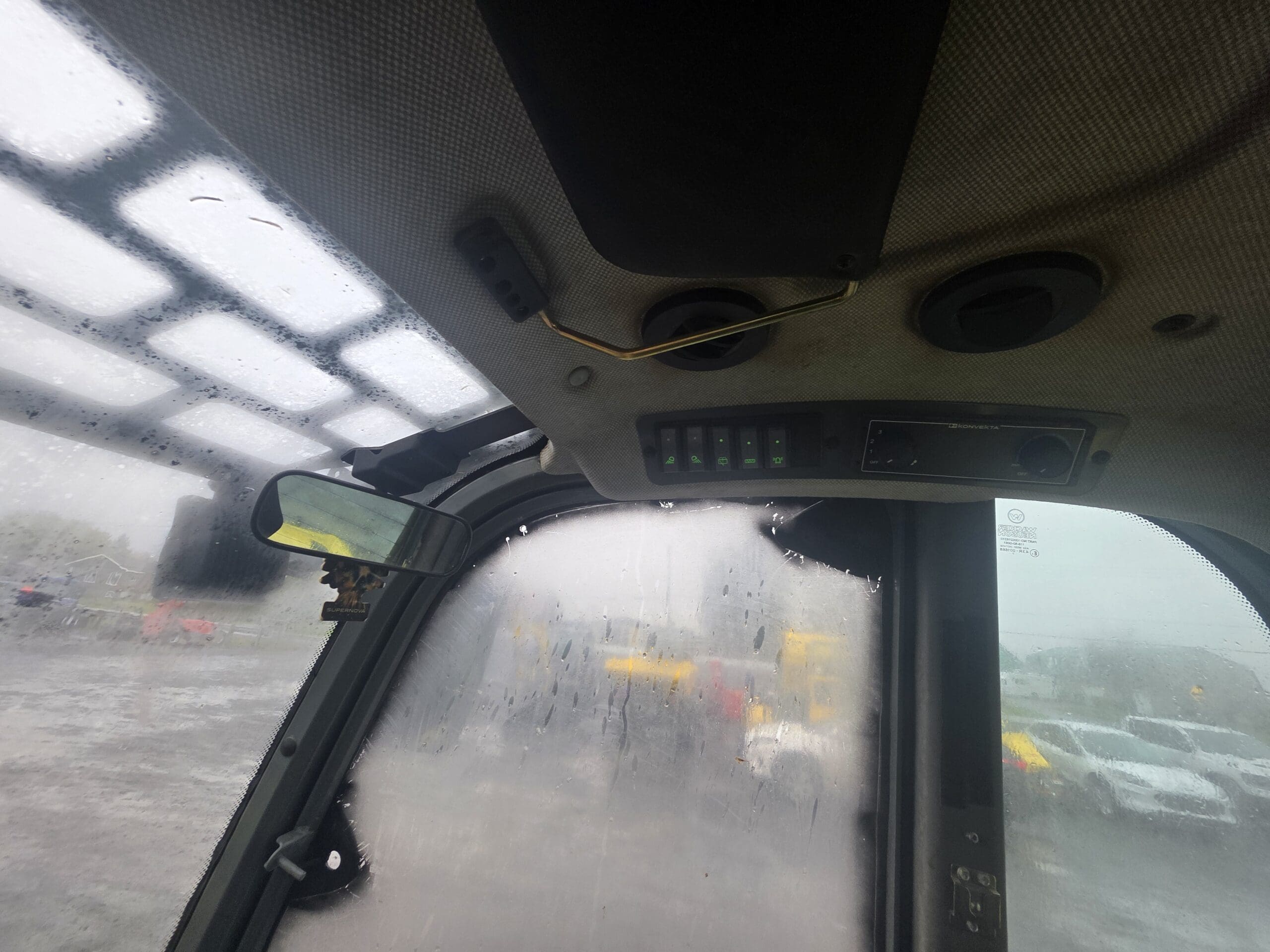 Vue depuis l'intérieur de la cabine d'un engin de chantier sous la pluie, montrant un tableau de commande fixé au plafond et des vitres embuées donnant sur un parking.