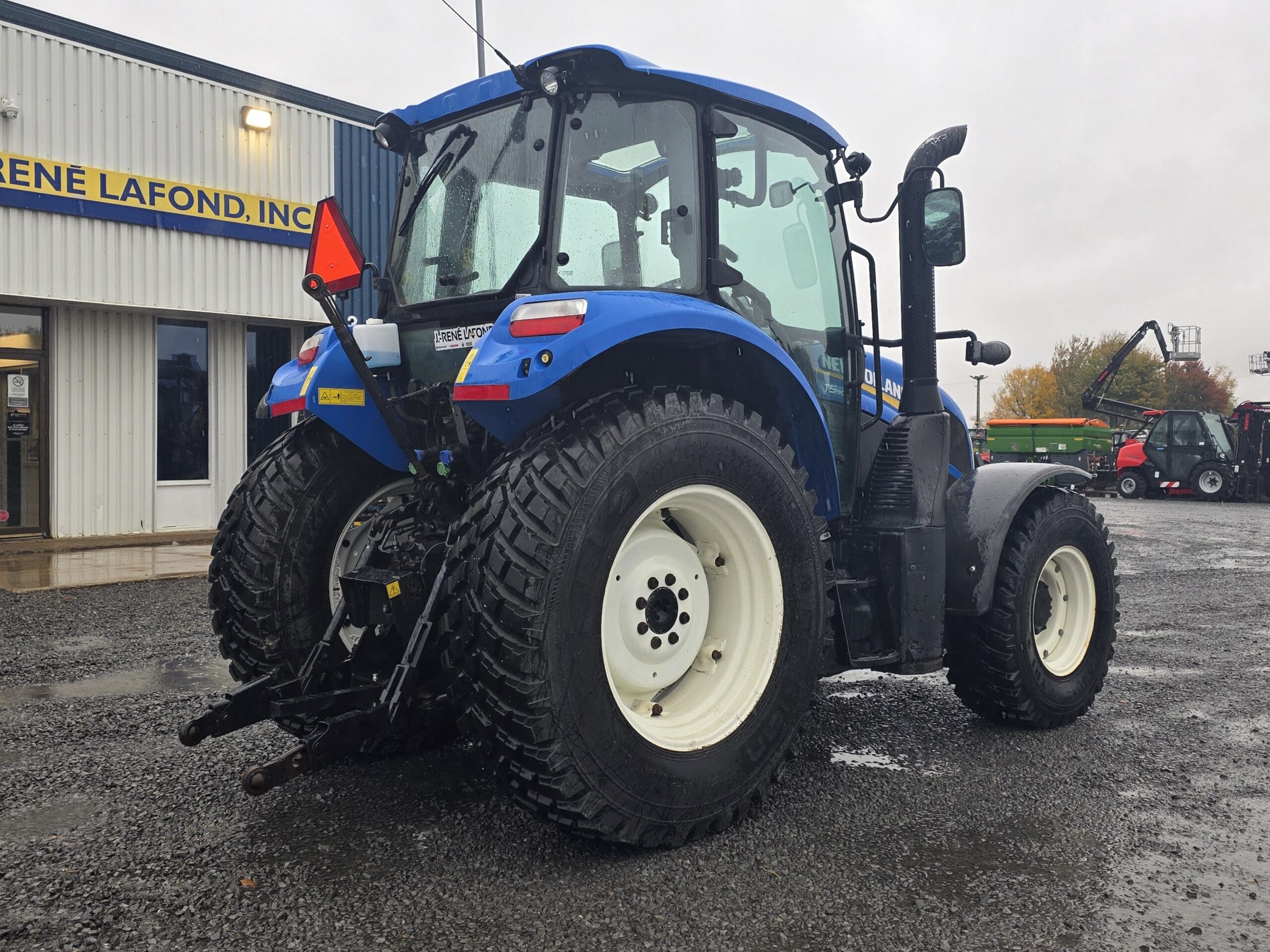 Vue arrière d'un tracteur New Holland bleu garé sur un parking en gravier devant un bâtiment.