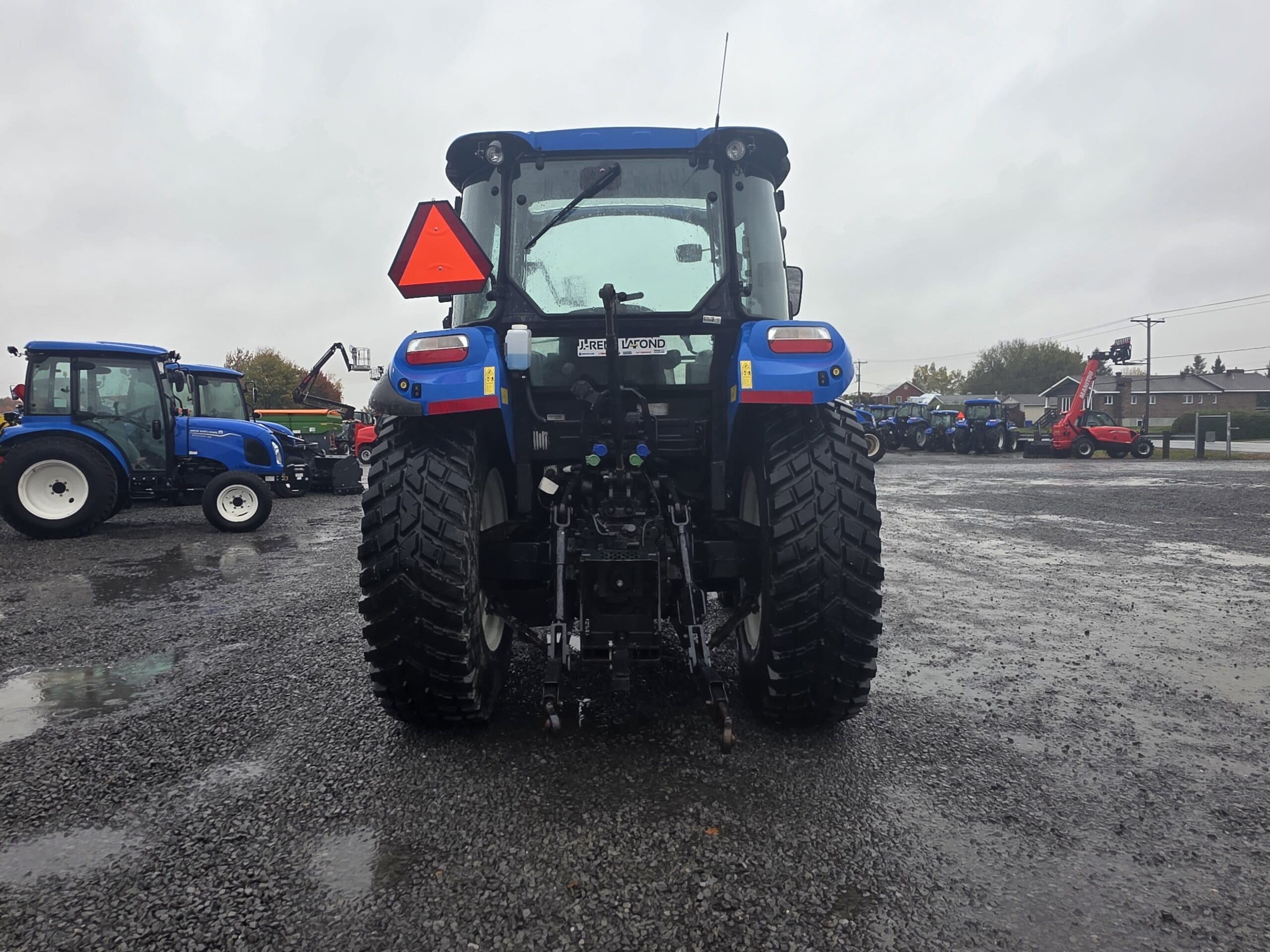 Vue arrière d'un tracteur bleu arborant un panneau triangulaire rouge signalant un véhicule lent, garé sur un terrain en gravier avec d'autres tracteurs en arrière-plan.