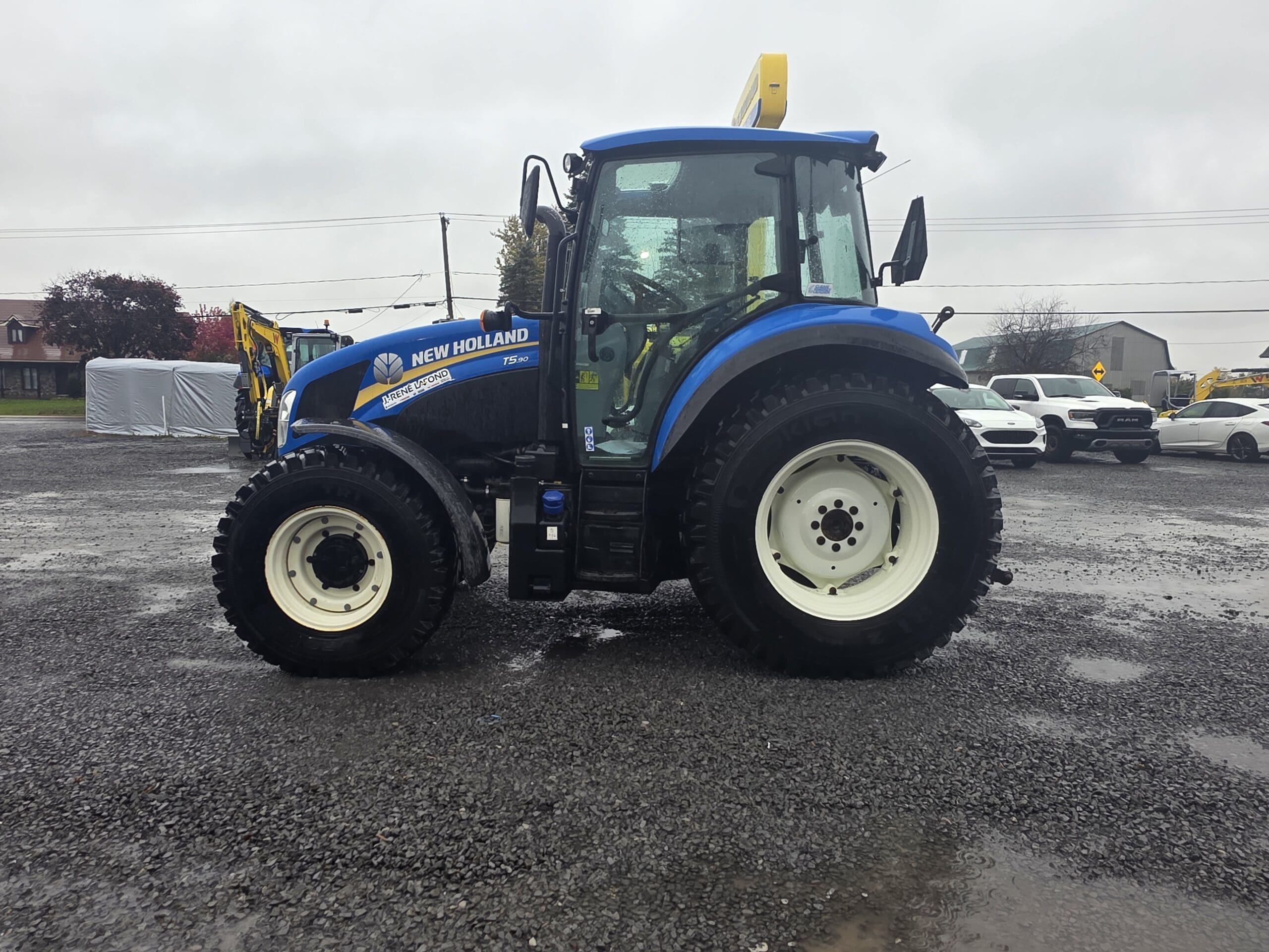 Un tracteur New Holland T5.80 bleu garé sur un terrain en gravier détrempé.