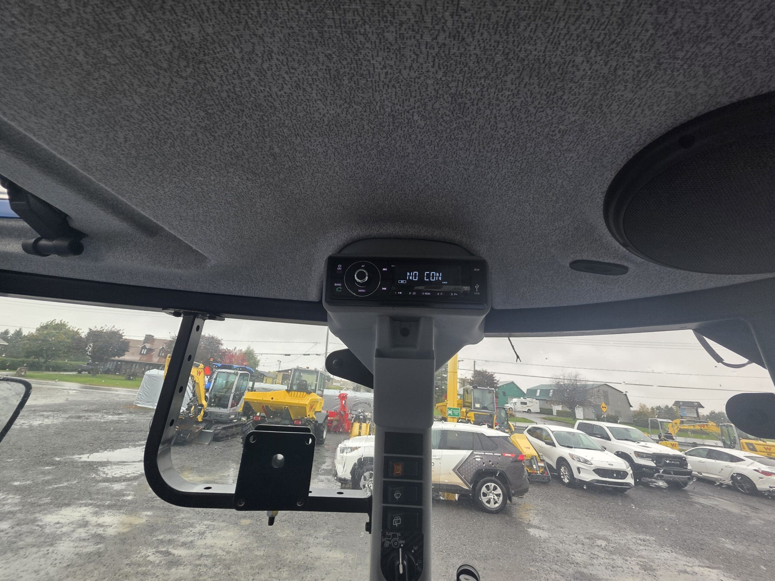 Vue depuis l'intérieur de la cabine d'un engin de chantier, donnant sur un parking rempli de voitures de toutes sortes et d'engins de chantier jaunes.