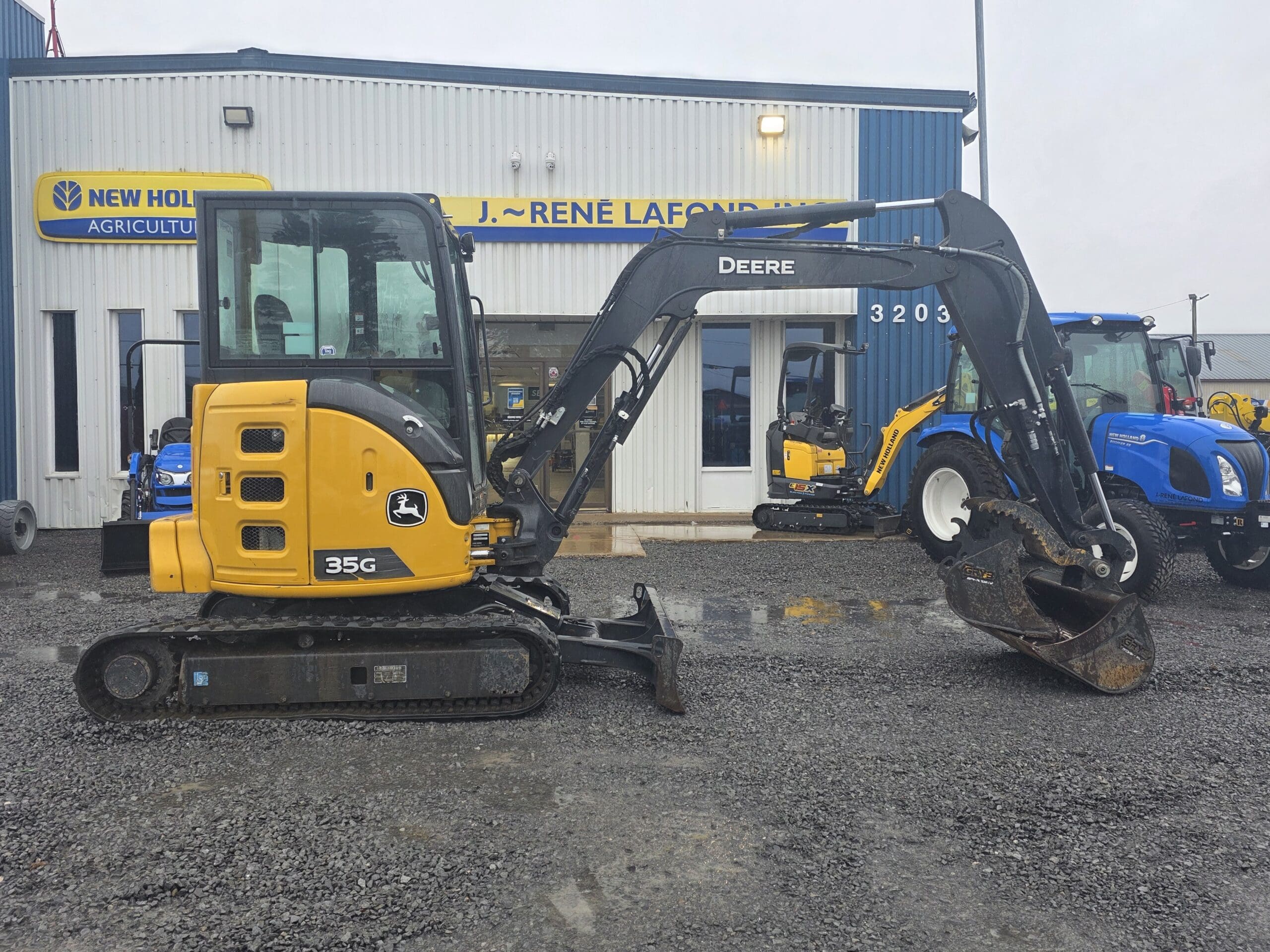 Une petite pelleteuse John Deere 35G jaune garée devant un concessionnaire agricole New Holland.