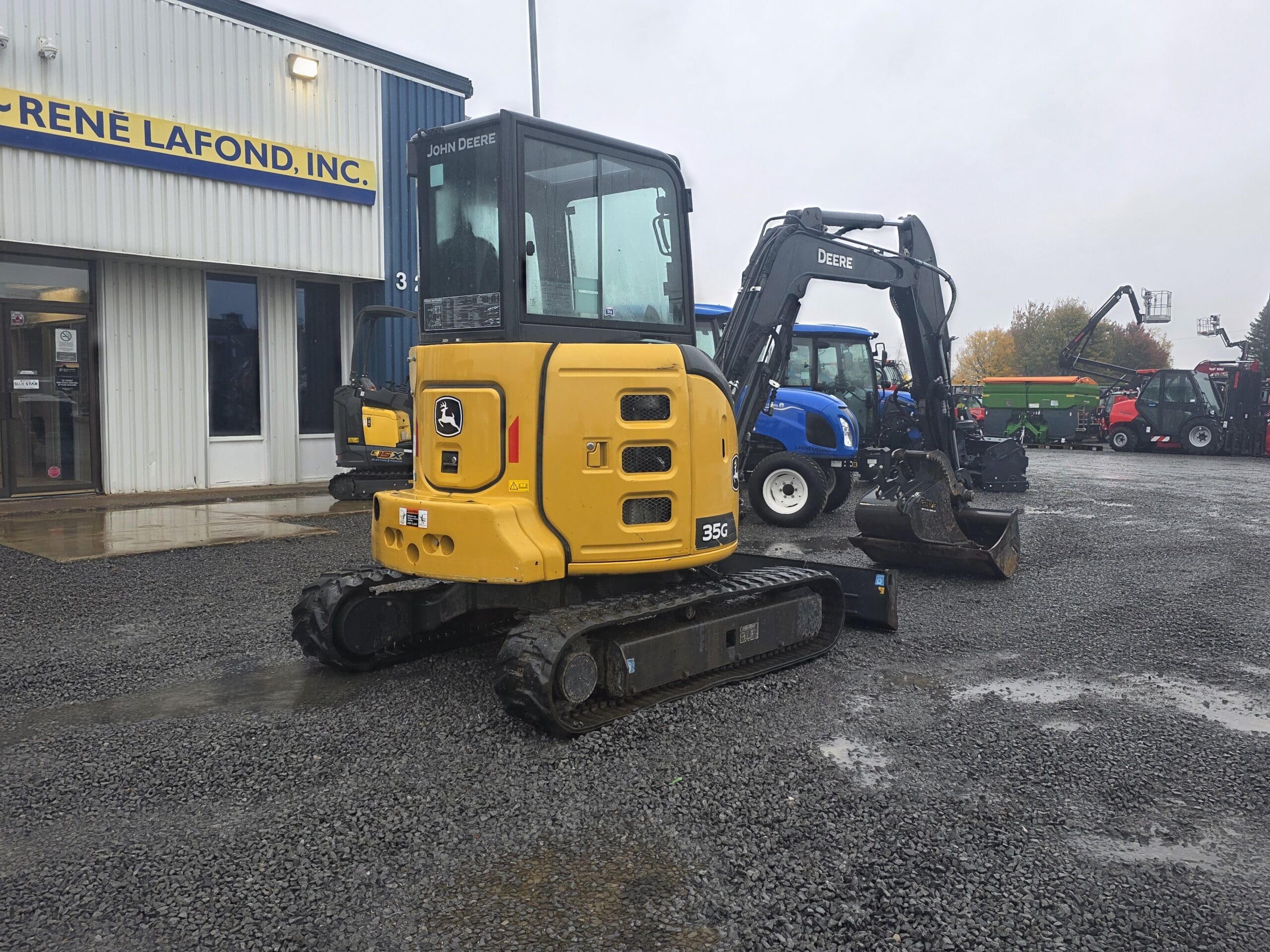 Une pelle compacte John Deere 35G jaune, garée sur un terrain en gravier devant un bâtiment.