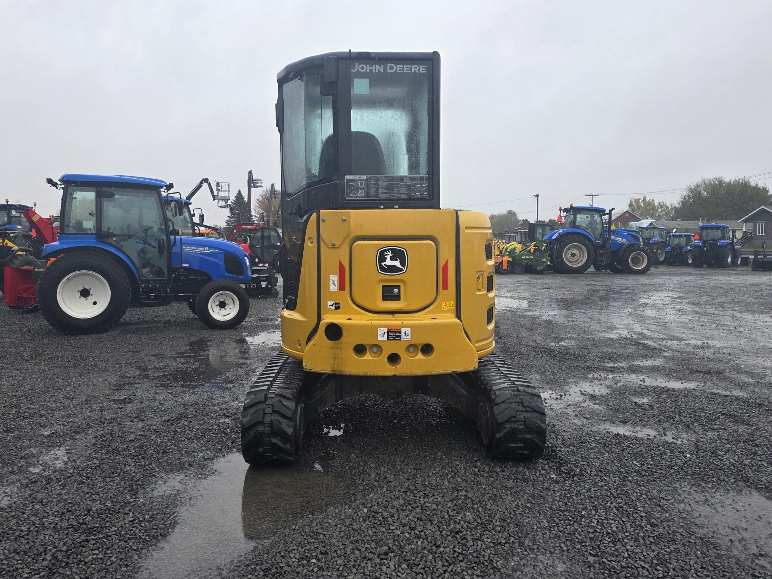 Une pelleteuse compacte John Deere jaune garée sur un terrain en gravier, entourée de tracteurs bleus.