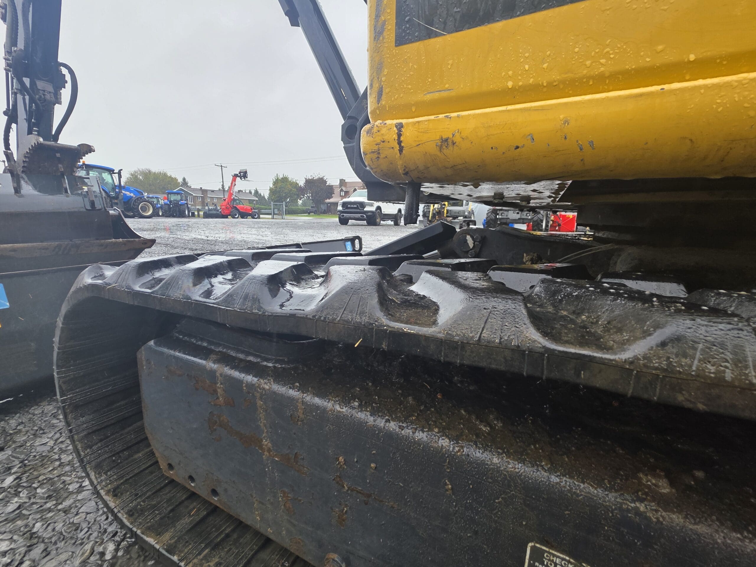 Gros plan sur les chenilles en caoutchouc noir et la carrosserie jaune d'une pelleteuse à l'arrêt, avec d'autres engins de chantier et une camionnette visibles à l'arrière-plan.