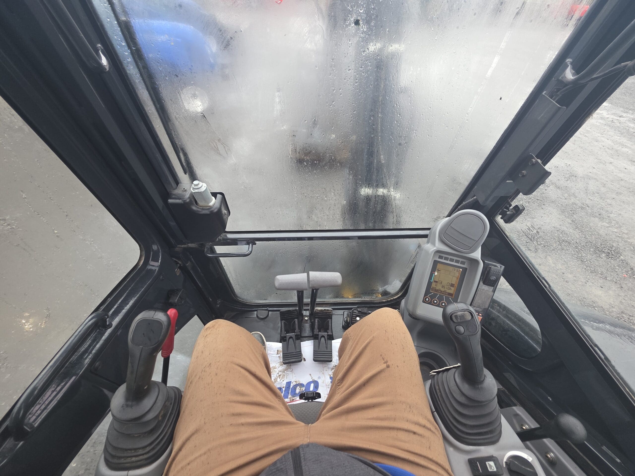 Vue à la première personne depuis la cabine d'une pelleteuse, montrant les jambes du conducteur et les manettes de commande à travers un pare-brise strié de gouttes de pluie.