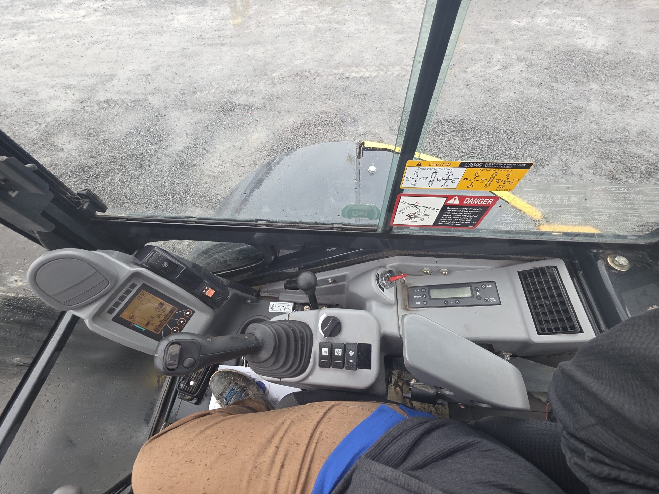 Vue depuis l'intérieur de la cabine d'une pelleteuse, montrant le joystick, les panneaux de commande et les autocollants de sécurité.