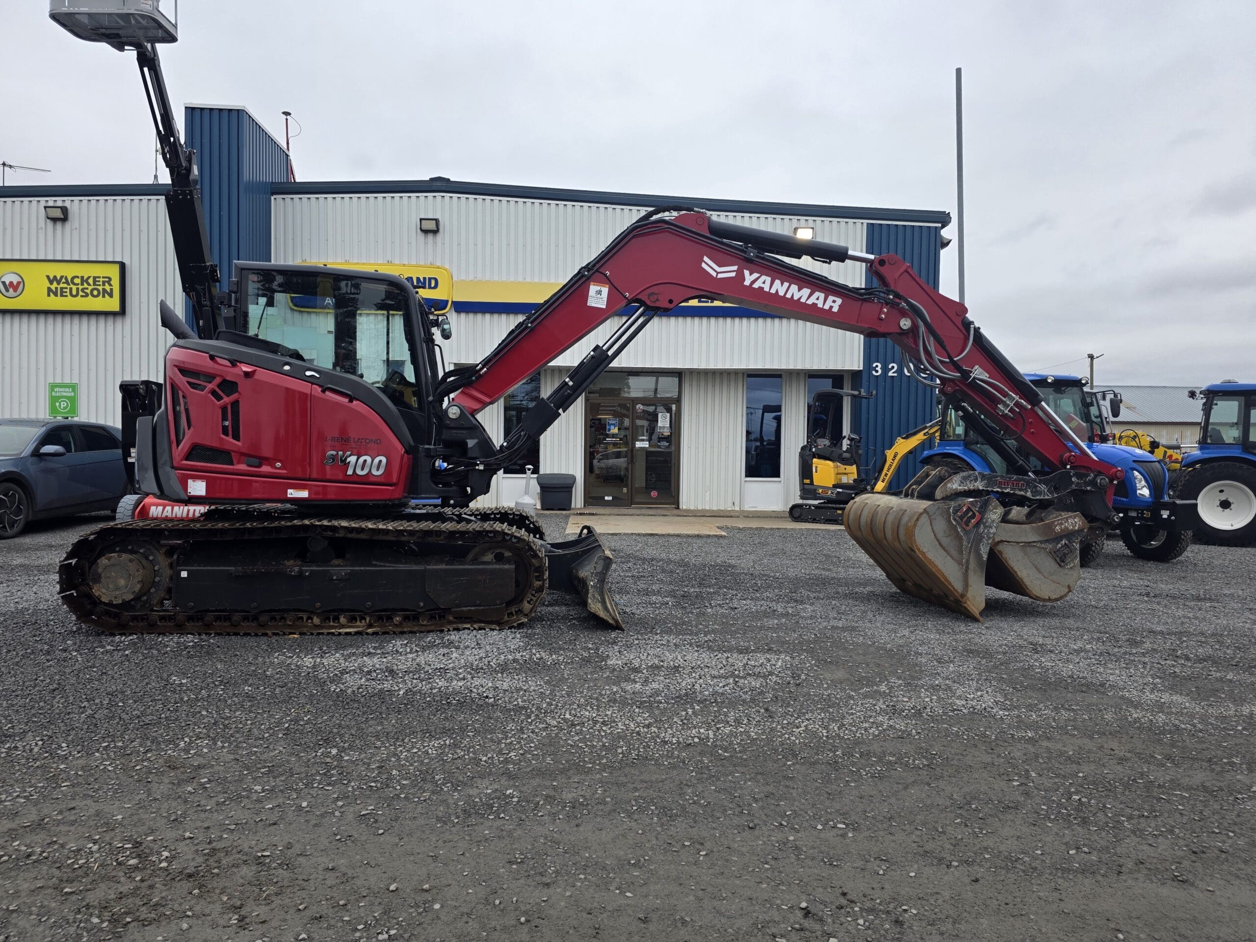 Une pelleteuse Yanmar SV100 rouge garée sur un parking en gravier devant une concession.