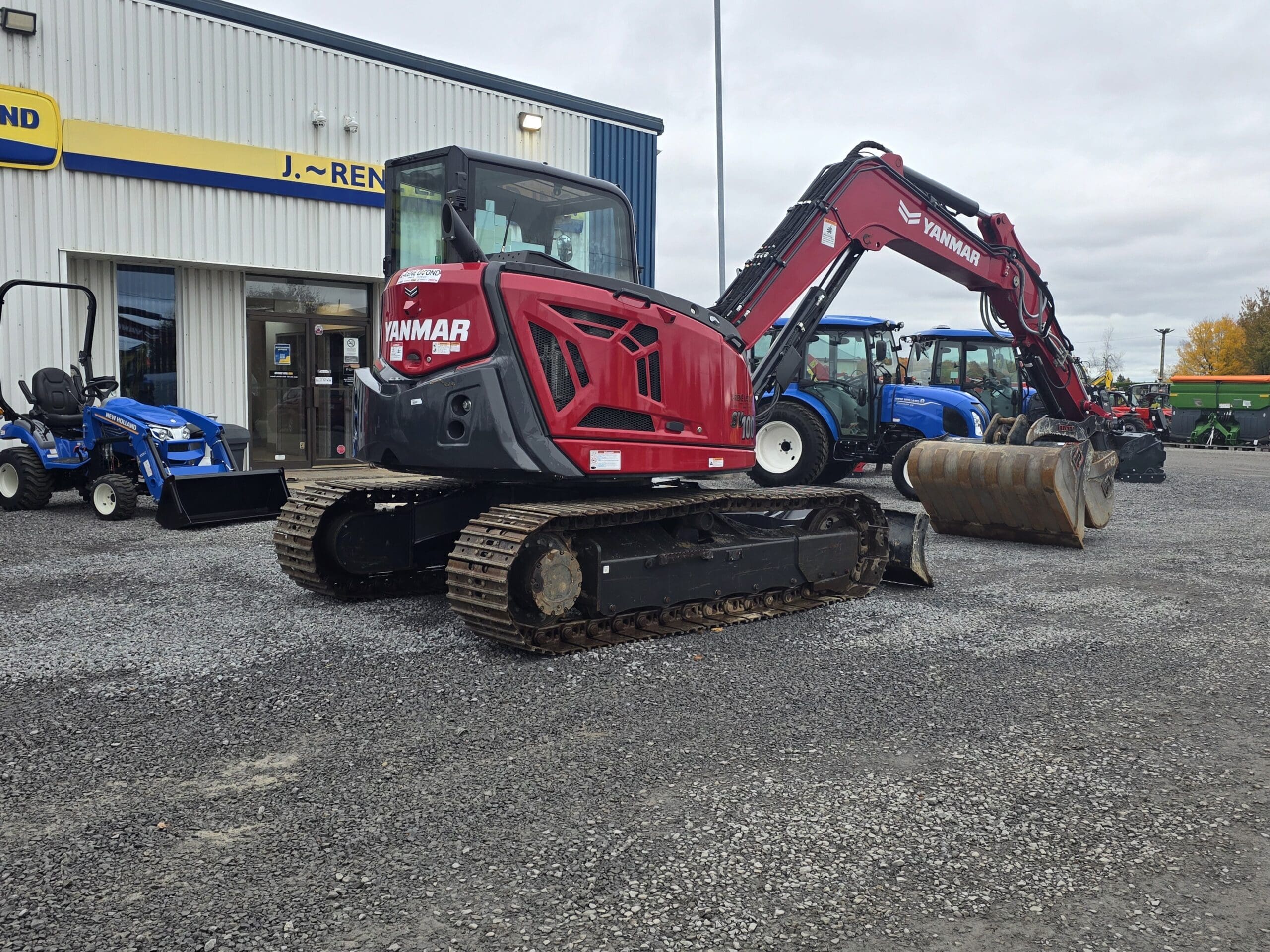 Une pelleteuse à chenilles Yanmar rouge garée sur un terrain en gravier devant un concessionnaire de machines.