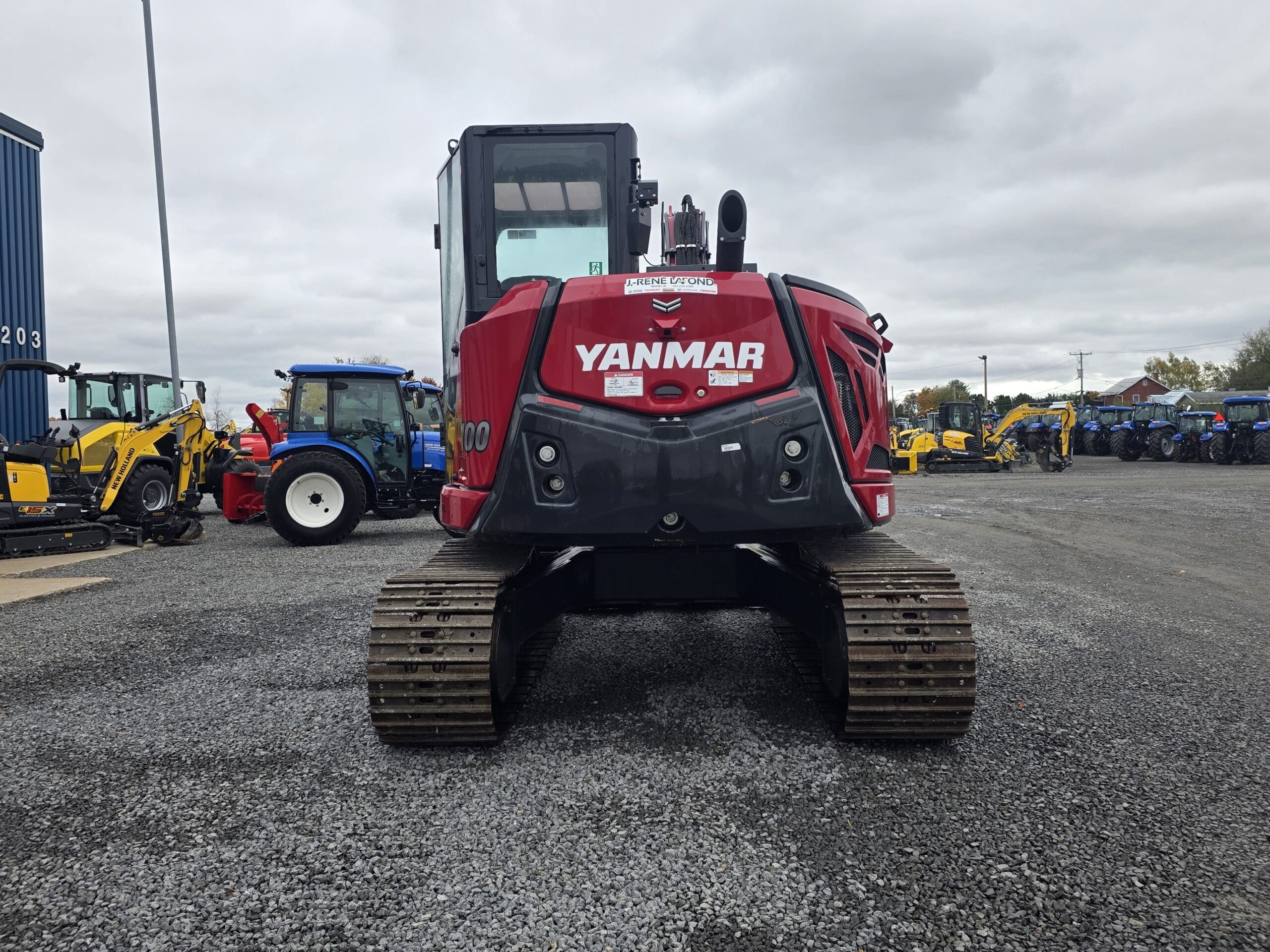 Une pelleteuse à chenilles Yanmar rouge garée sur un terrain en gravier, avec d'autres tracteurs et engins de chantier visibles à l'arrière-plan.